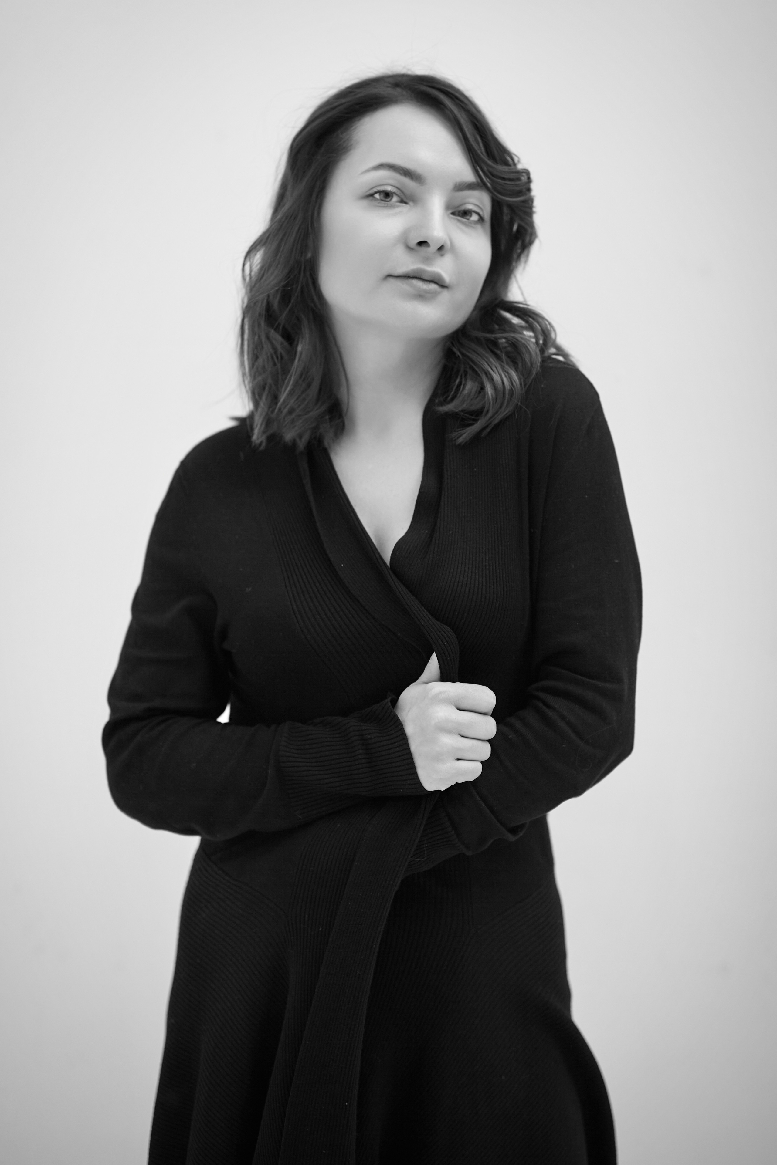 Темноволосая модель Татьяна позирует в светлом платье на стуле на фоне светлой стены в студии.