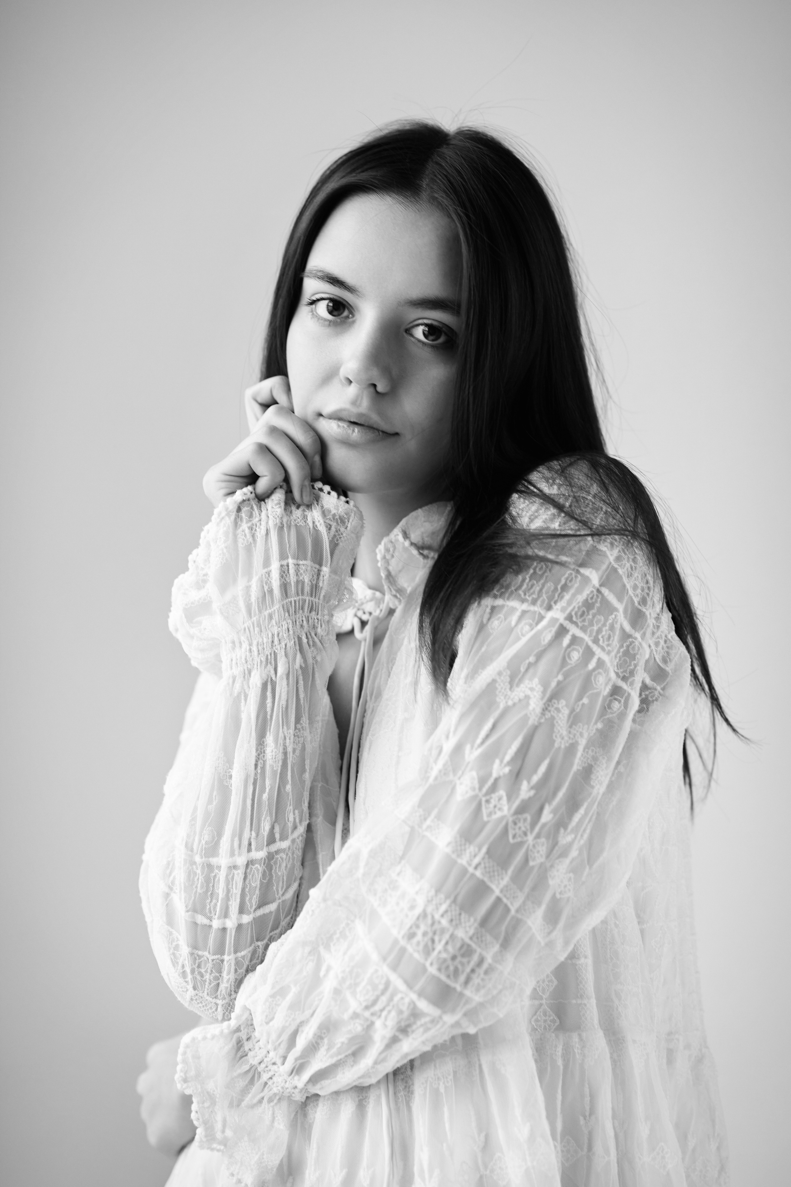 Темноволосая Амина на светлом фоне в студии одета в светлое платье, ее глаза светятся ярко.