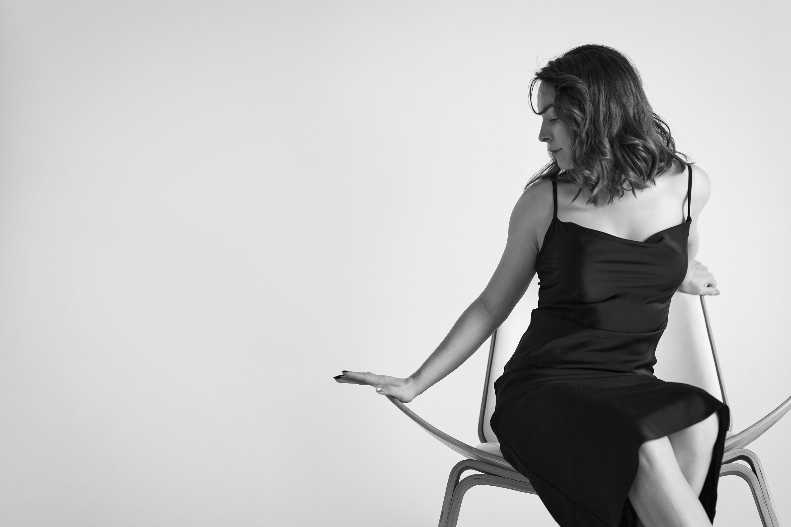 Татьяна, модель с темными волосами, на светлом фоне, в светлом платье, рубашке или топике, сидя на стуле с небольшой улыбкой.