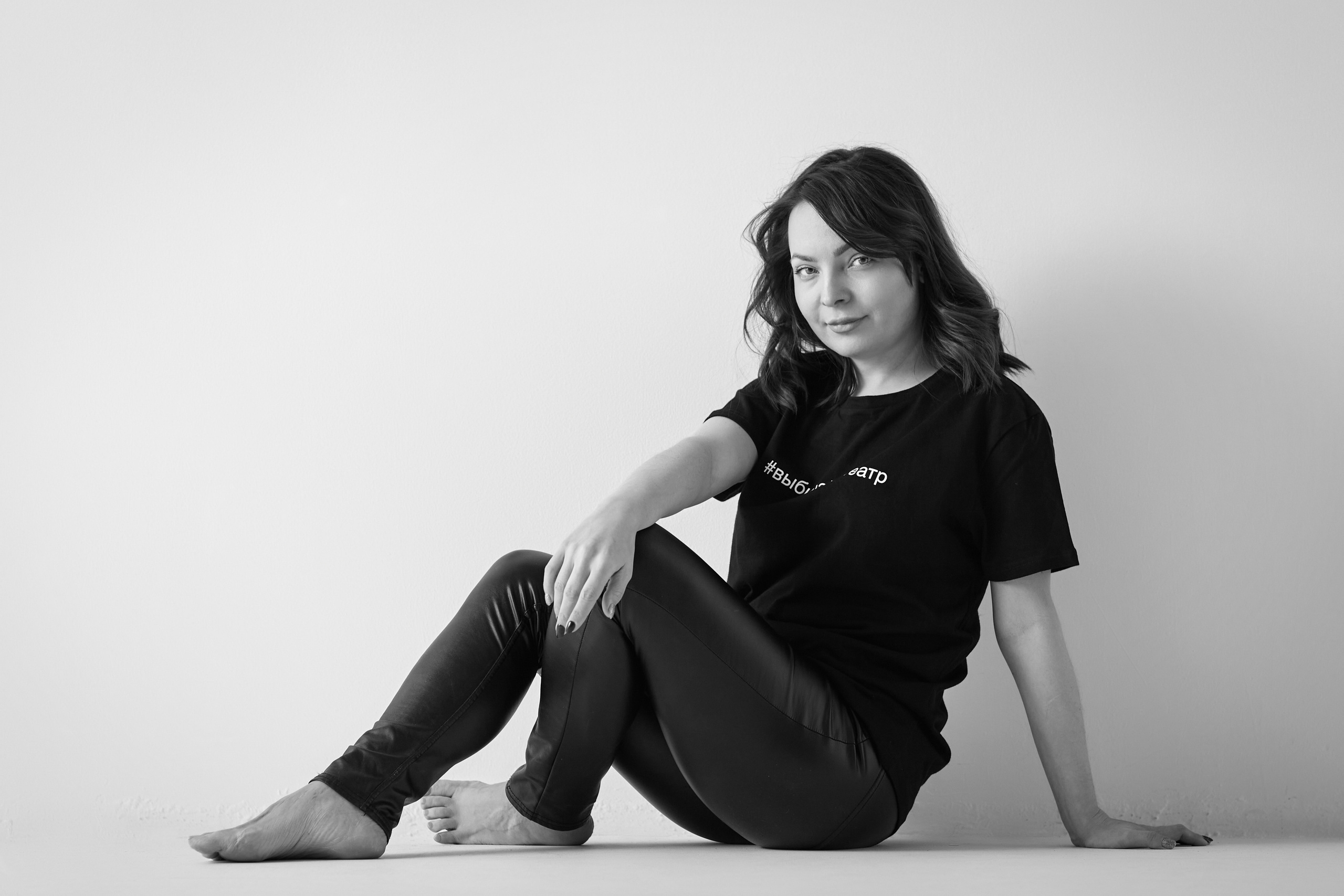 Темноволосая модель Татьяна на светлом фоне в светлом платье или в рубашке или в топике или на стуле, смотрящая в камеру с легкой улыбкой.