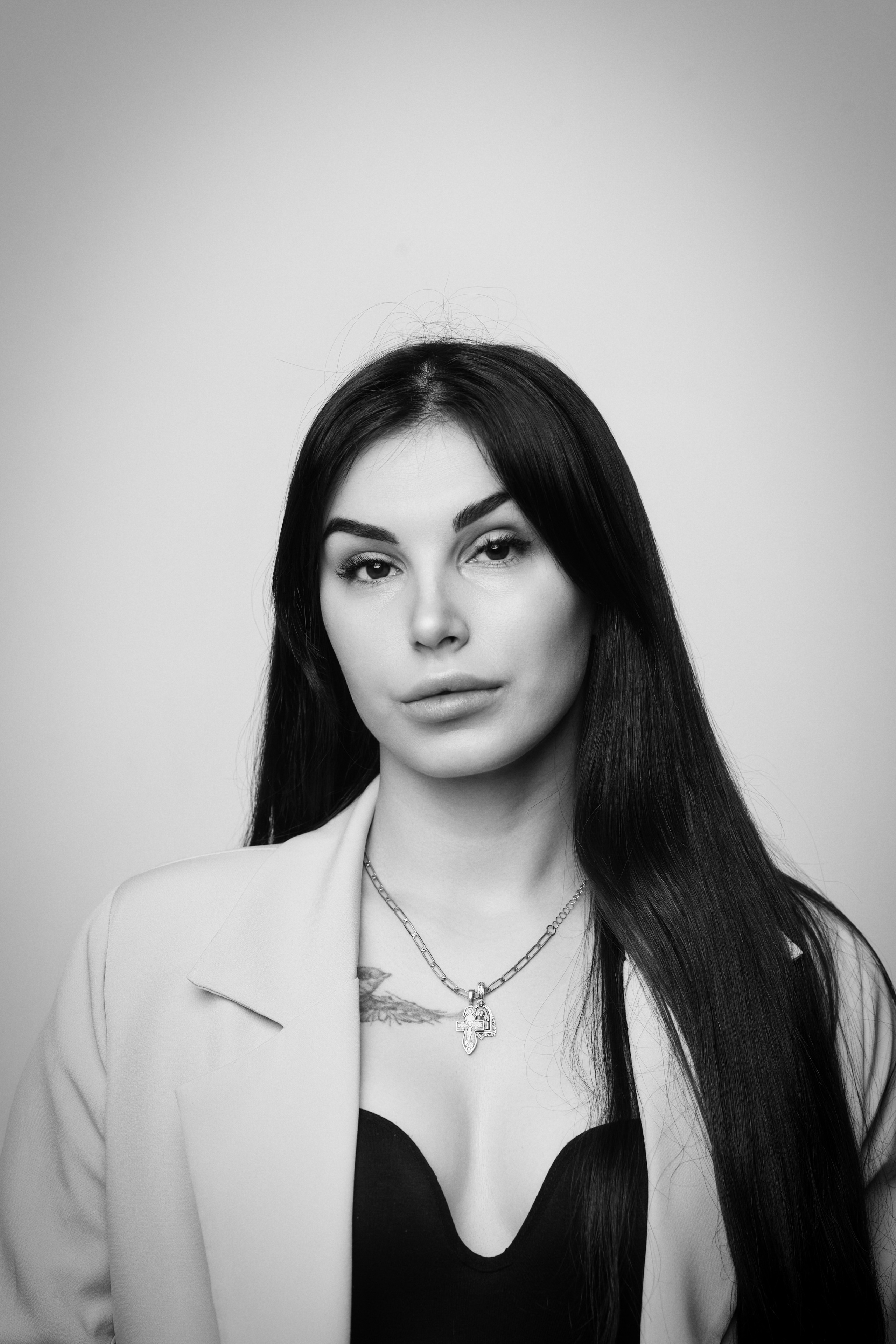 В студии на светлом фоне темноволосая Карина в рубашке позирует перед камерой, демонстрируя свою красоту.