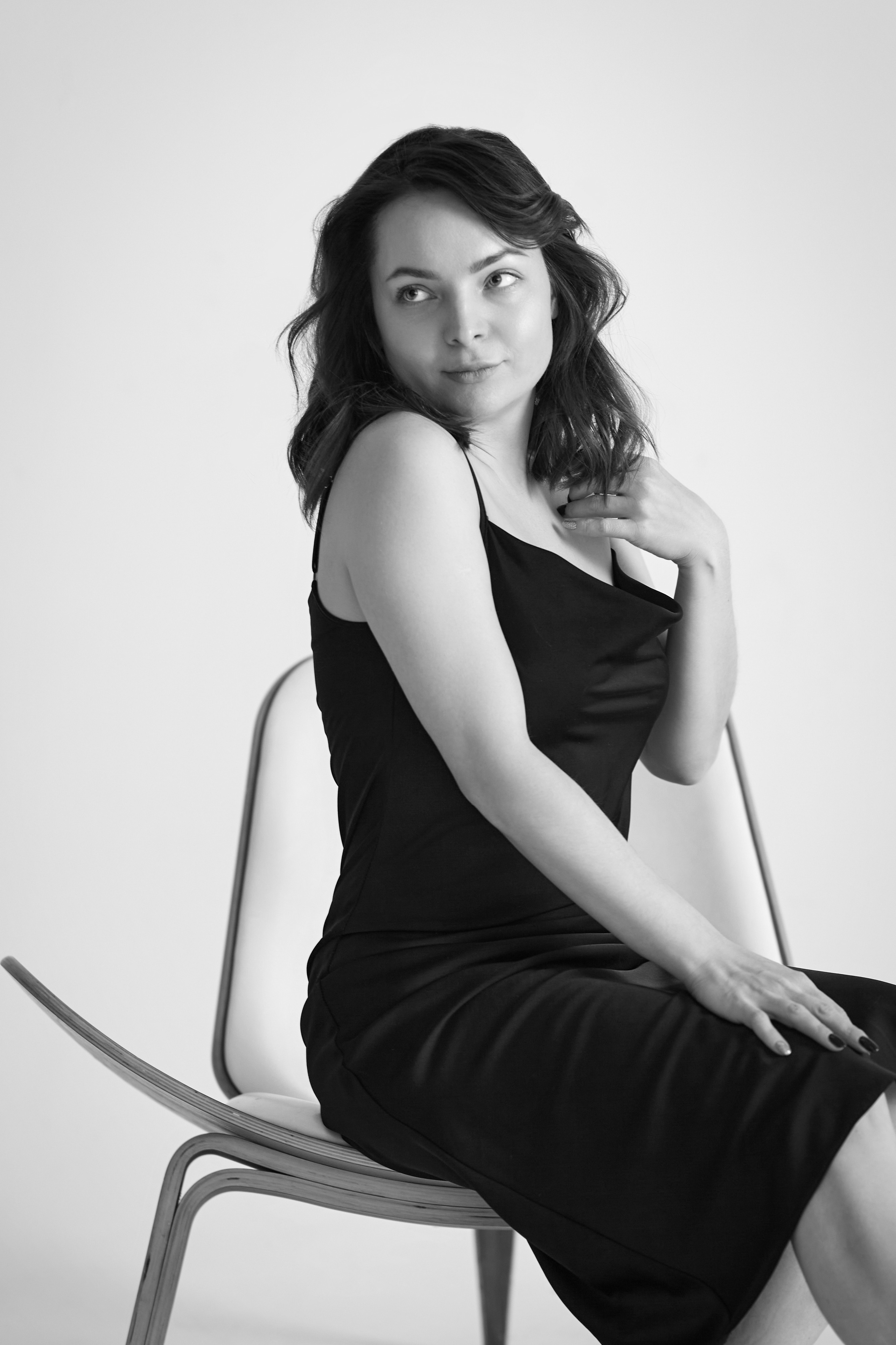 Темноволосая модель Татьяна на светлом фоне, в светлом платье, рубашке или топике, сидящая на стуле и смотрящая в камеру.