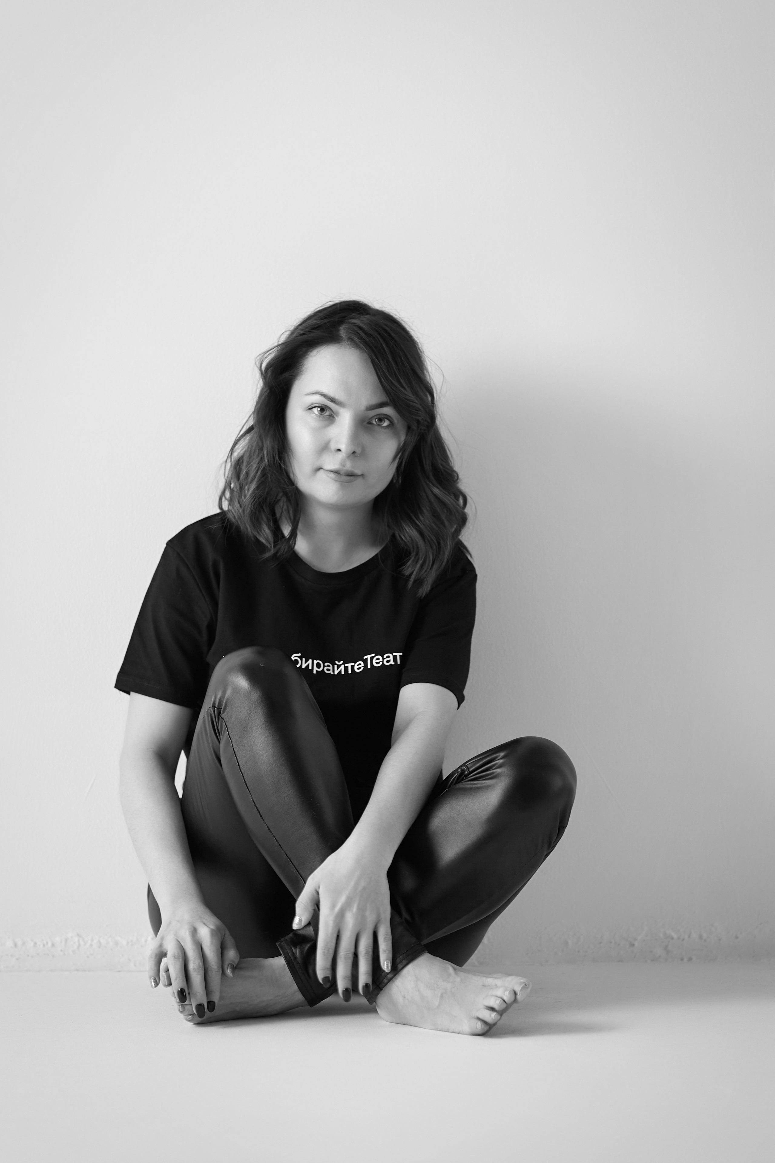 Темноволосая модель Татьяна сидит на стуле в светлом топике на фоне светлой стены в студии.