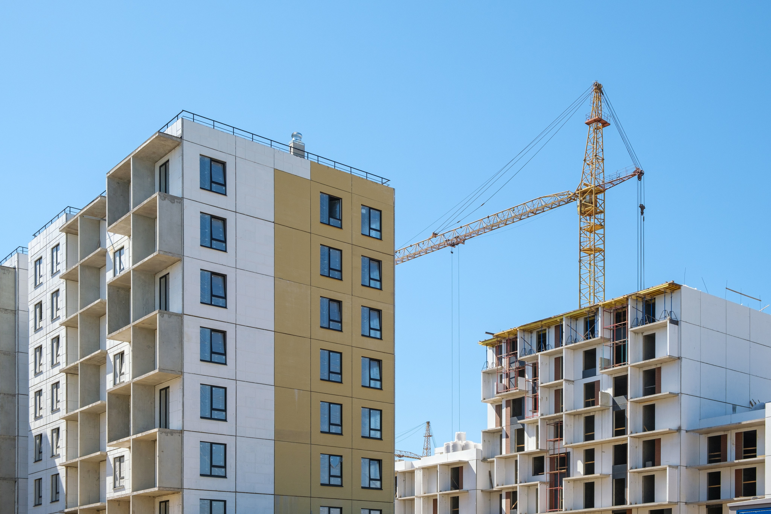 На изображении представлены два строящихся многоквартирных дома разной этажности. Слева находится здание с большим количеством окон и несколькими балконами, окрашенное в светло-желтый цвет. Справа расположен дом с более простой архитектурой и меньшим количеством этажей, выполненный в белом цвете. На заднем плане виден строительный кран, что указывает на продолжающиеся строительные работы.