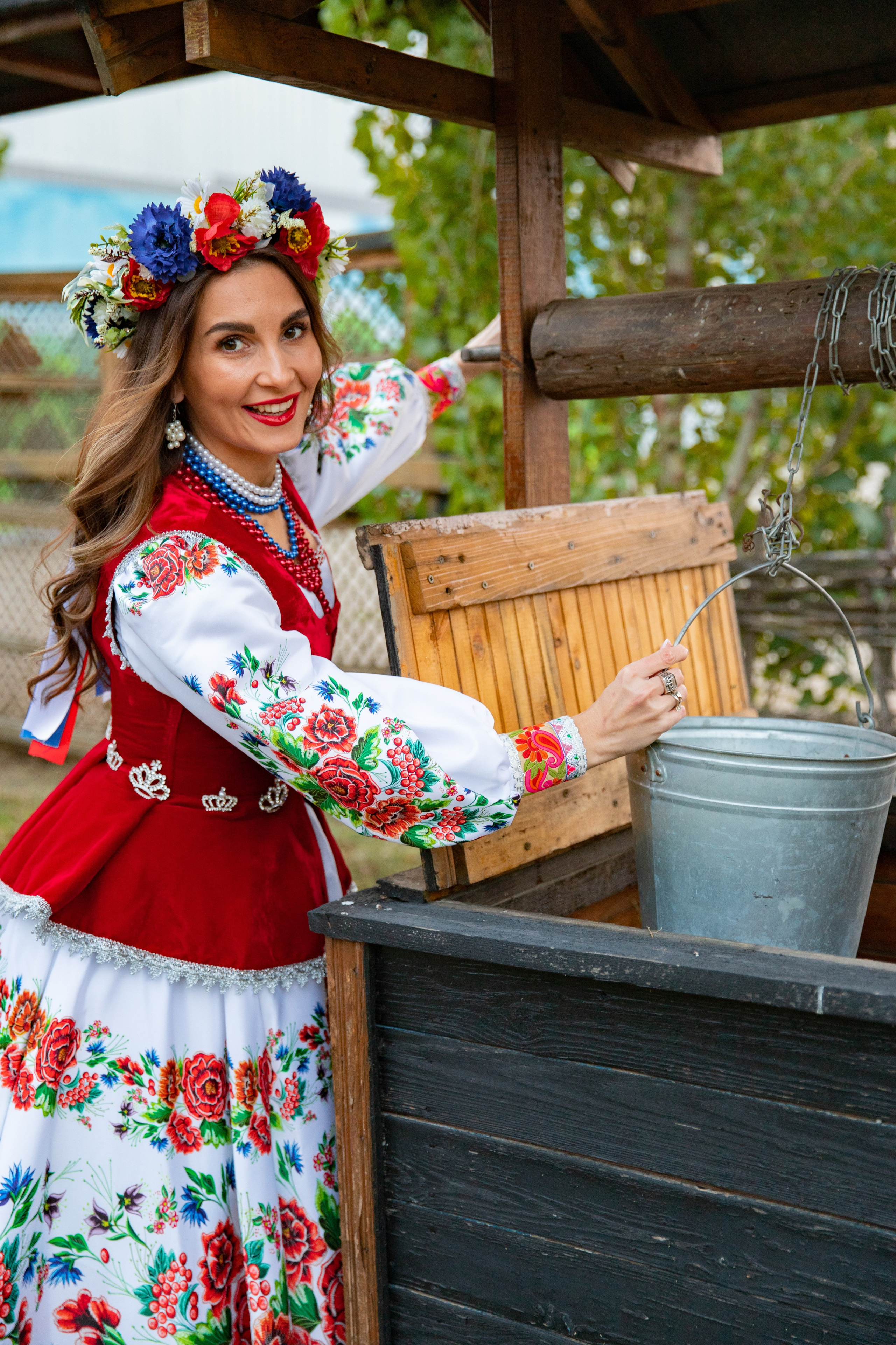 Девушка в красочном платье казачки стоит у старинного колодца, подчеркивая связь с природой и народными традициями.