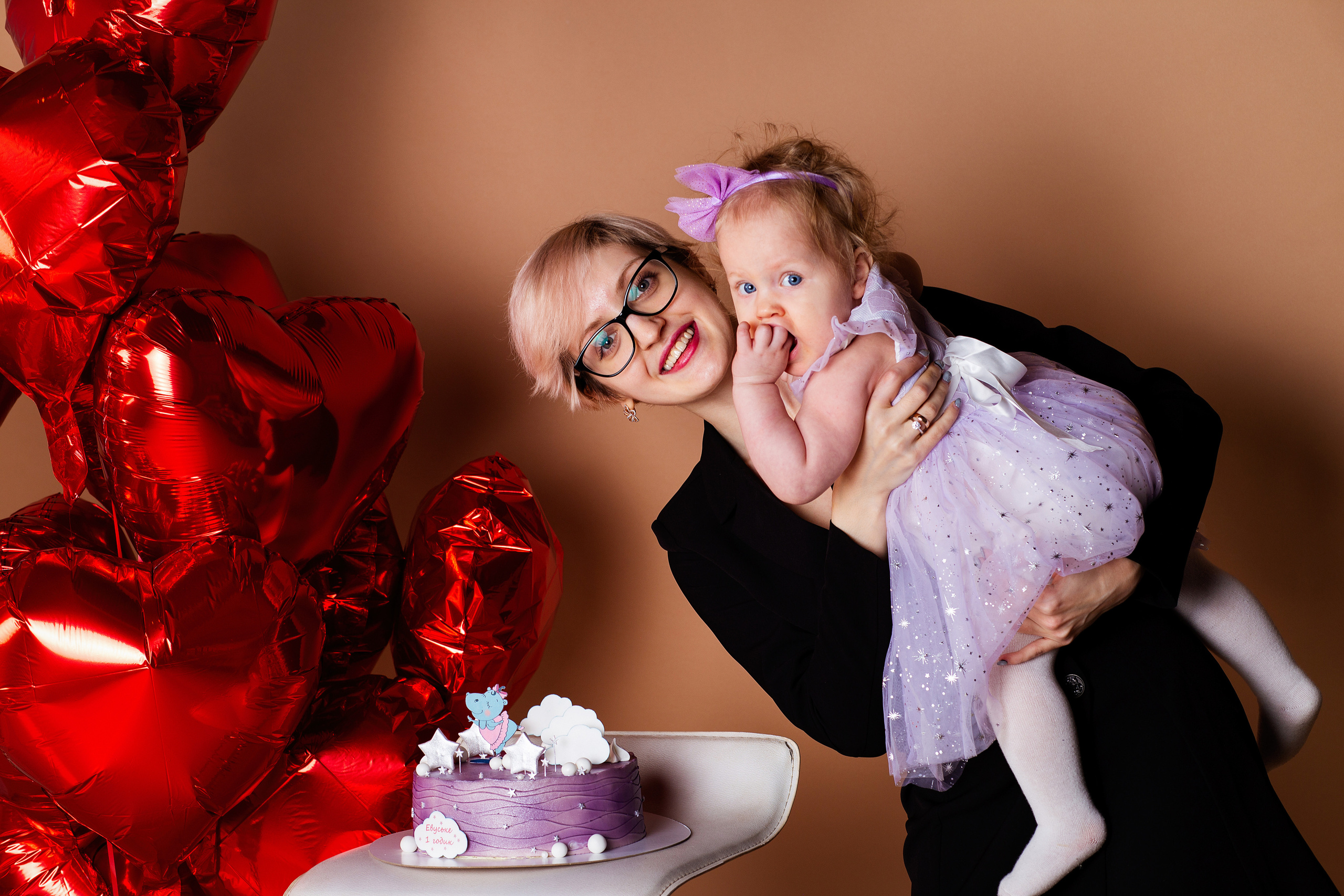 девушка  в очках  держит на  руках ребенка. рядом красные  шарики  и сиреневый торт