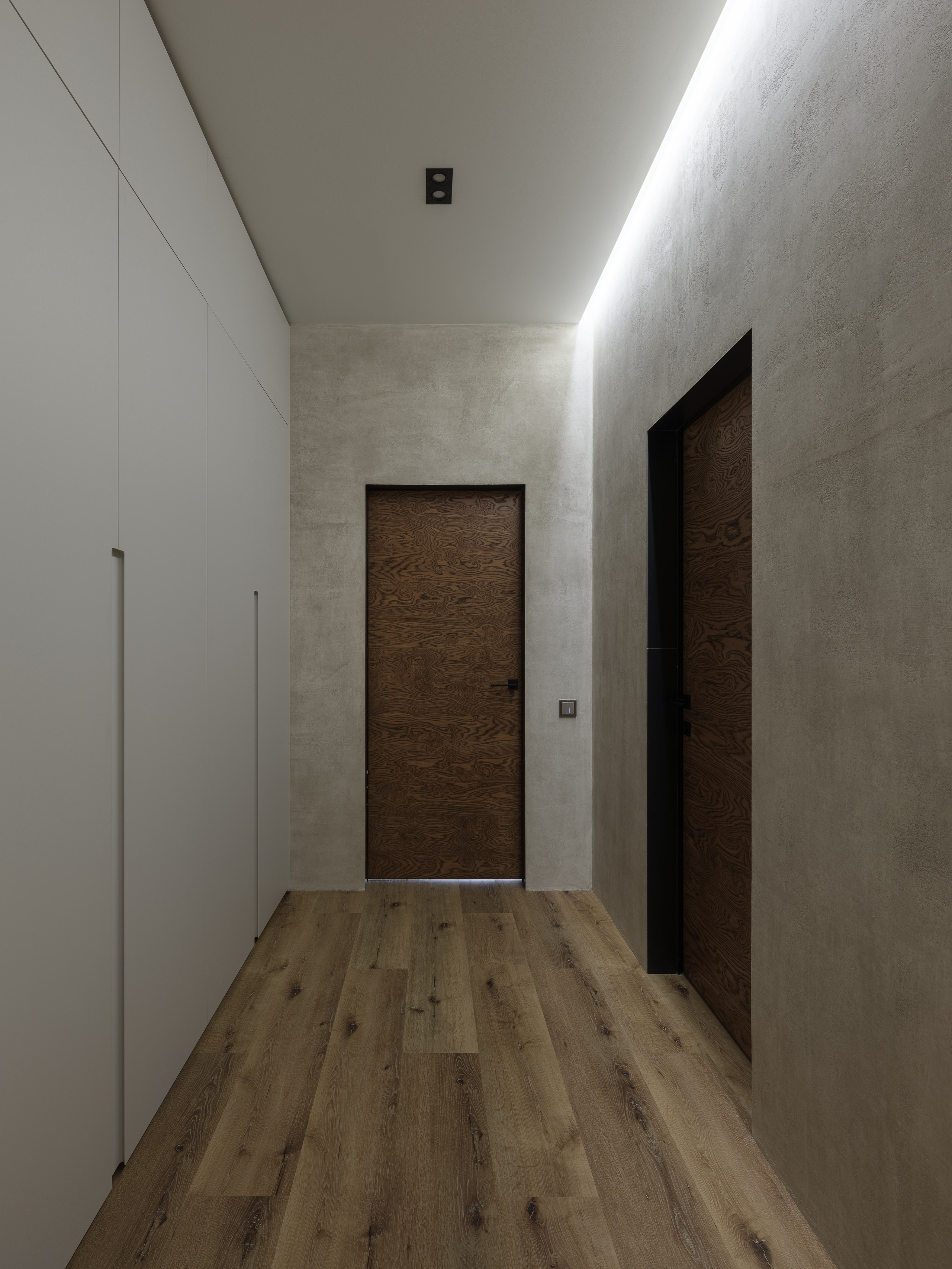 Минималистичный коридор с деревянным полом и дверями из темного шпона, светлыми стенами под бетон и встроенными шкафами без ручек; мягкая светодиодная подсветка создает атмосферу уюта. Интерьер оформлен дизайнером Алиной Дёровой, стилист Наталья Якушева, фото Сергей Денисенко.