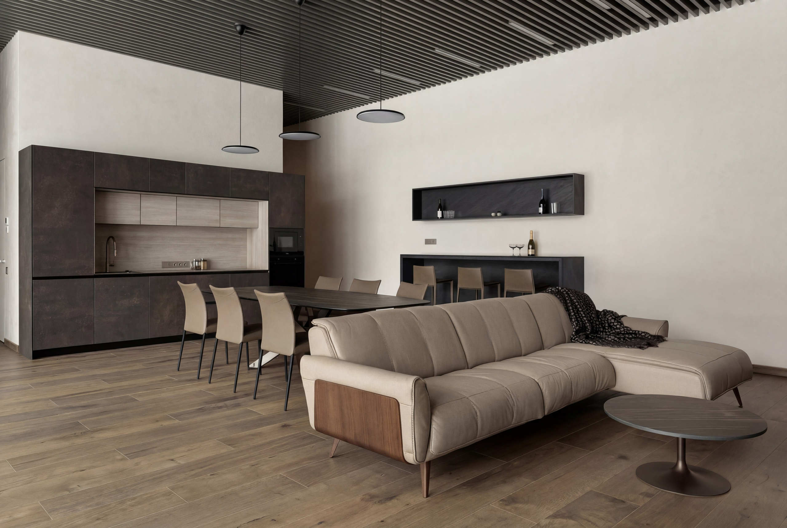 Интерьер lounge-зоны и темной кухни от Валерии Котович: кожаный угловой диван, обеденный стол с бежевыми креслами, реечный потолок и лаконичные черные светильники в современном доме у воды.