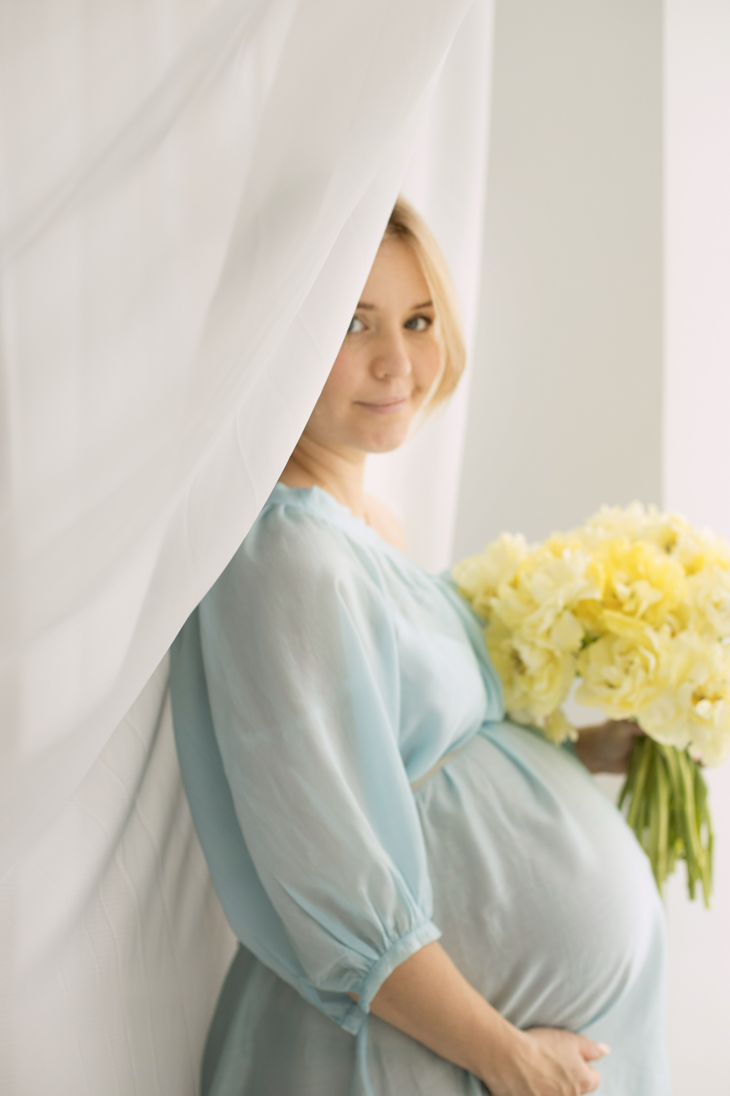 Беременная женщина в  голубом платье с букетом желтых пионов стоит у окна с белыми занавесками.