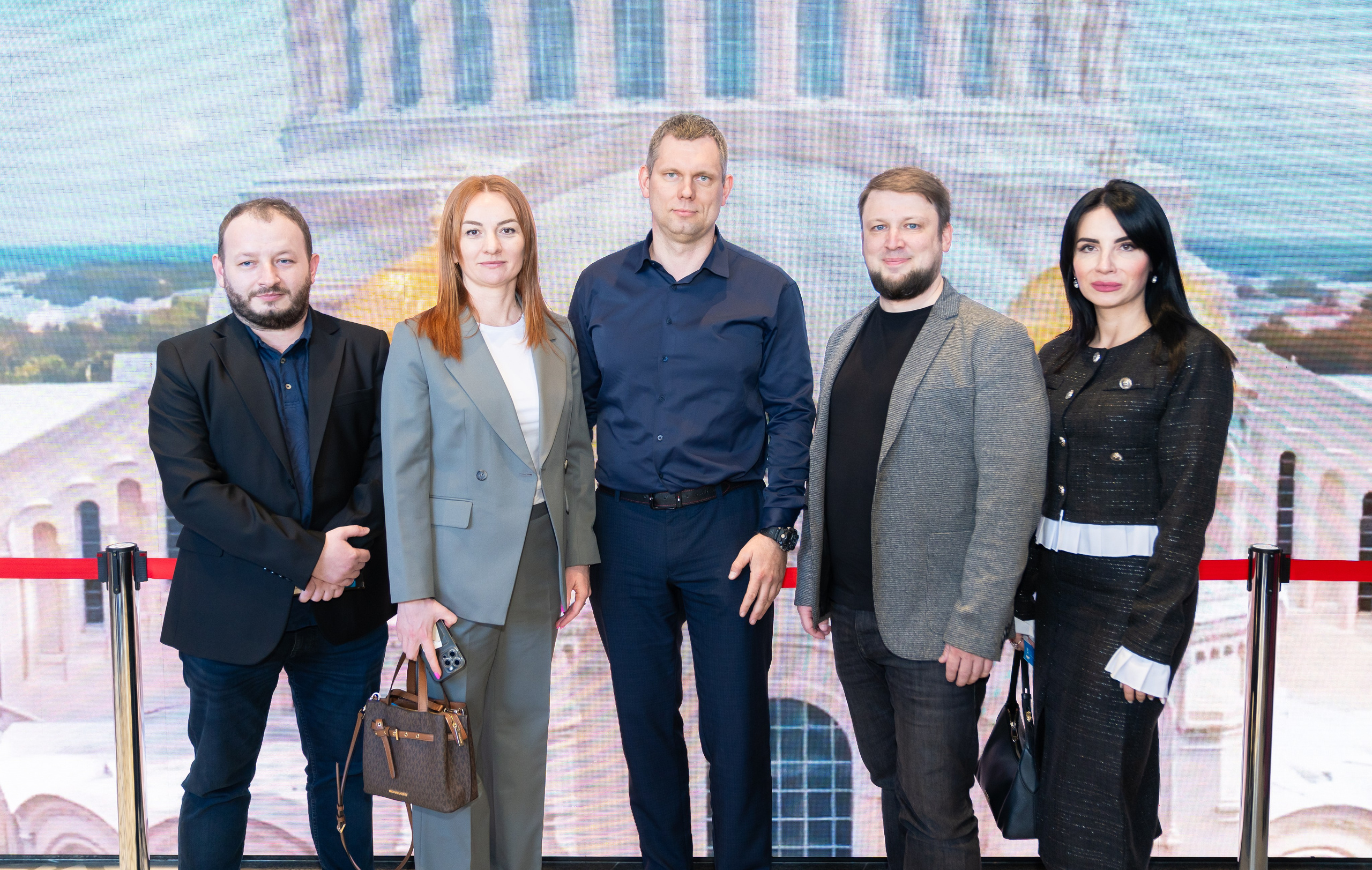 Групповое фото участников Дня местного самоуправления Санкт-Петербург. Пять человек в деловой одежде на фоне большой LED-панорамы города. Автор фото — Евгений Мальцев.