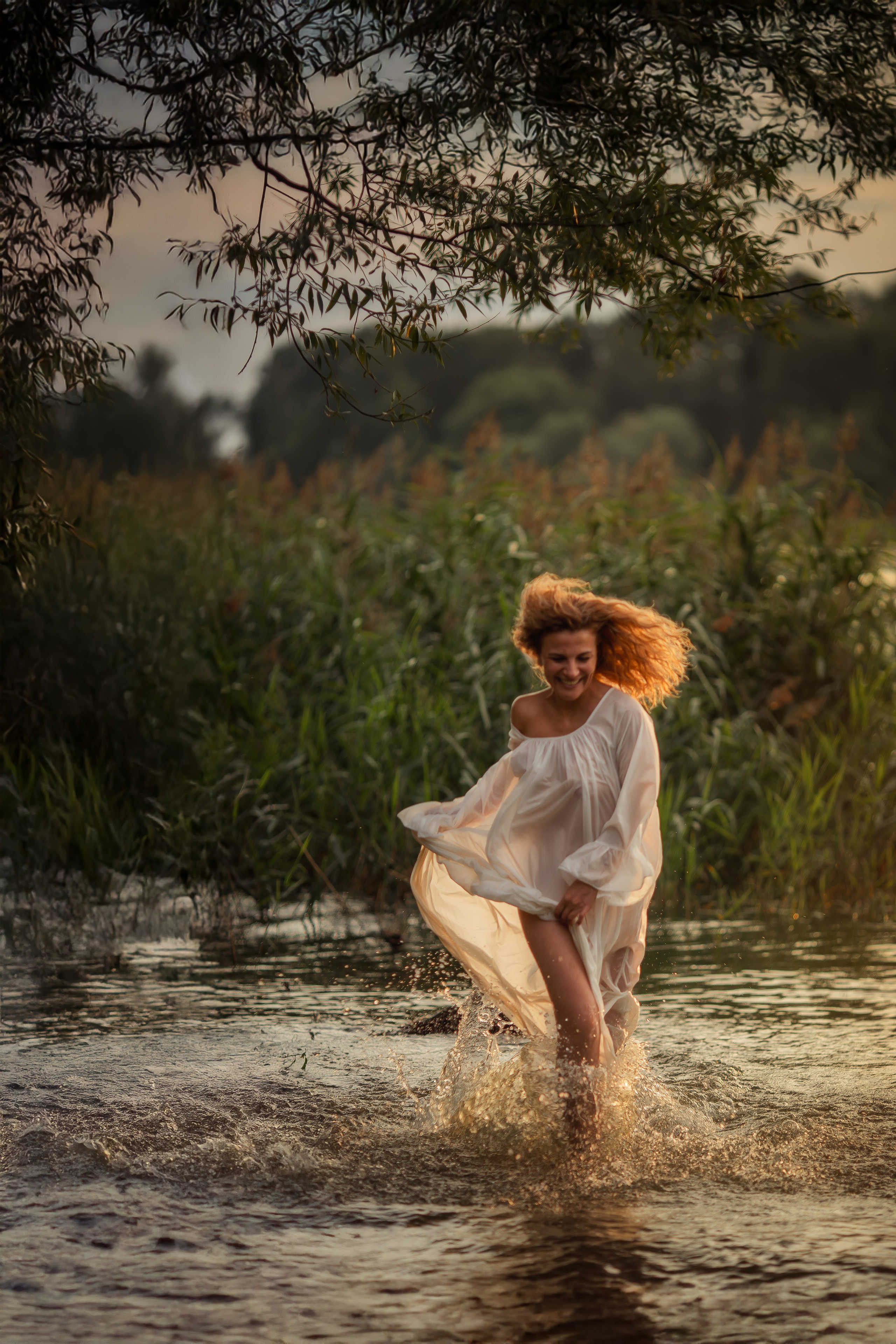 Девушка в белом воздушном платье бежит по воде, поднимая брызги, её волосы подсвечены золотым светом, на лице радостная улыбка, вокруг зелёный берег и ветви дерева.