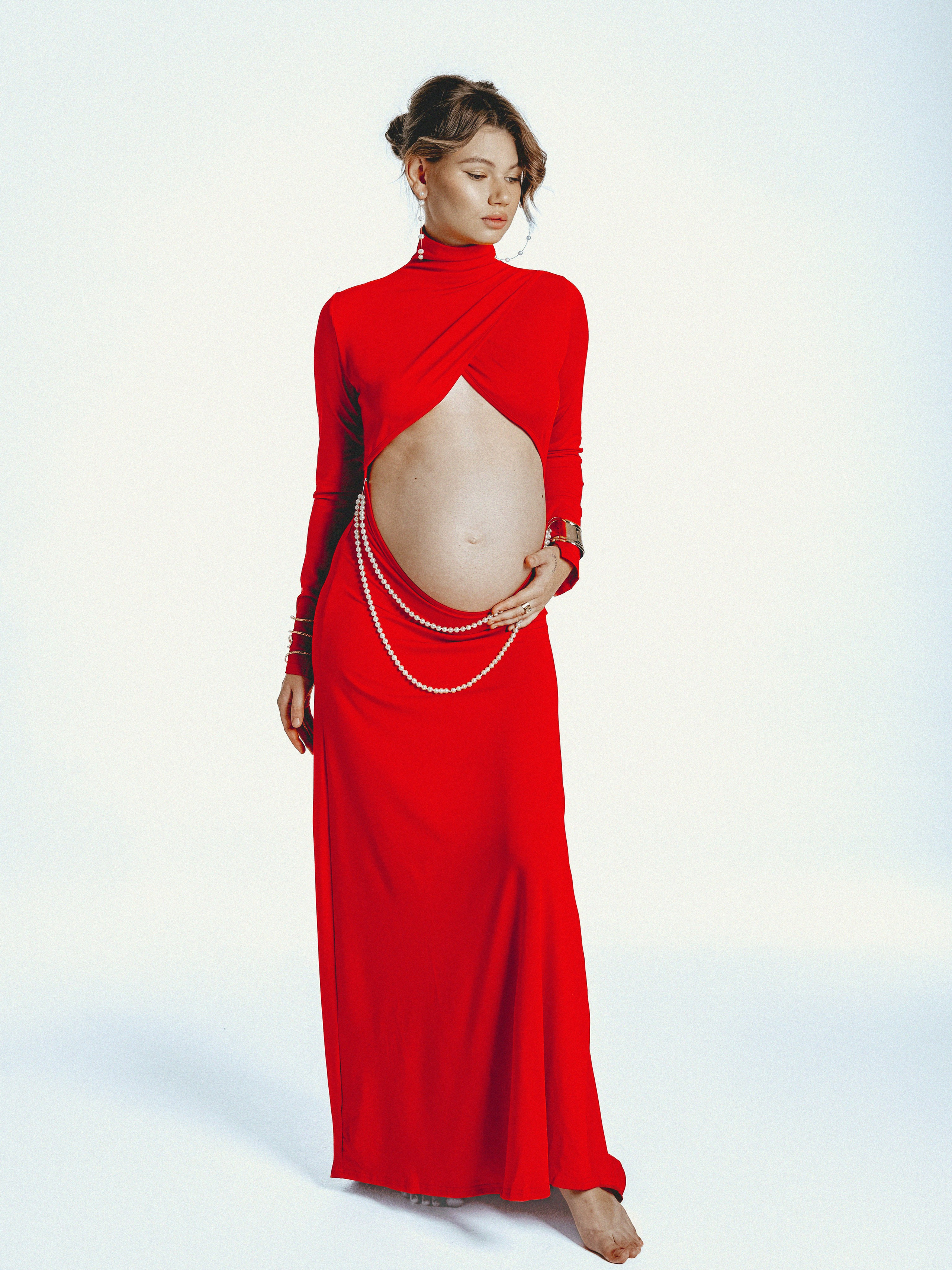 Беременная женщина в красном платье с жемчужным украшением на животе — стильная фотосессия для будущих мам в студии.