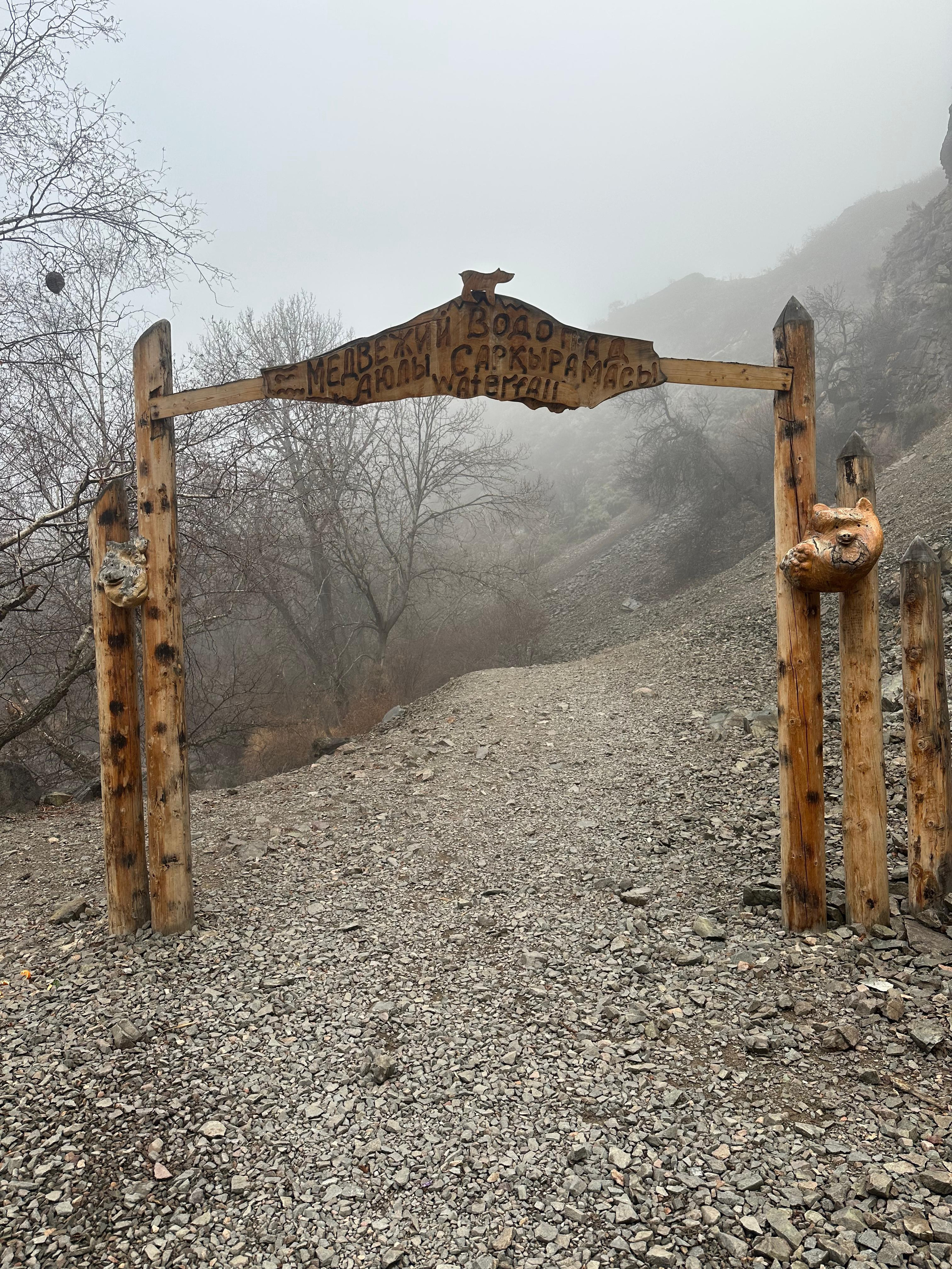 Деревянные ворота на входе в туристический маршрут, ведущий к водопаду Медвежий. На табличке, прикрепленной к воротам, написано: "Медвежий водопад" на нескольких языках, включая русский, казахский и английский ("Waterfall"). Сами ворота украшены резными деревянными элементами в виде медведя и медвежьей головы. Вокруг — туманная атмосфера с безлистными деревьями и каменистой тропой, ведущей вверх по склону.