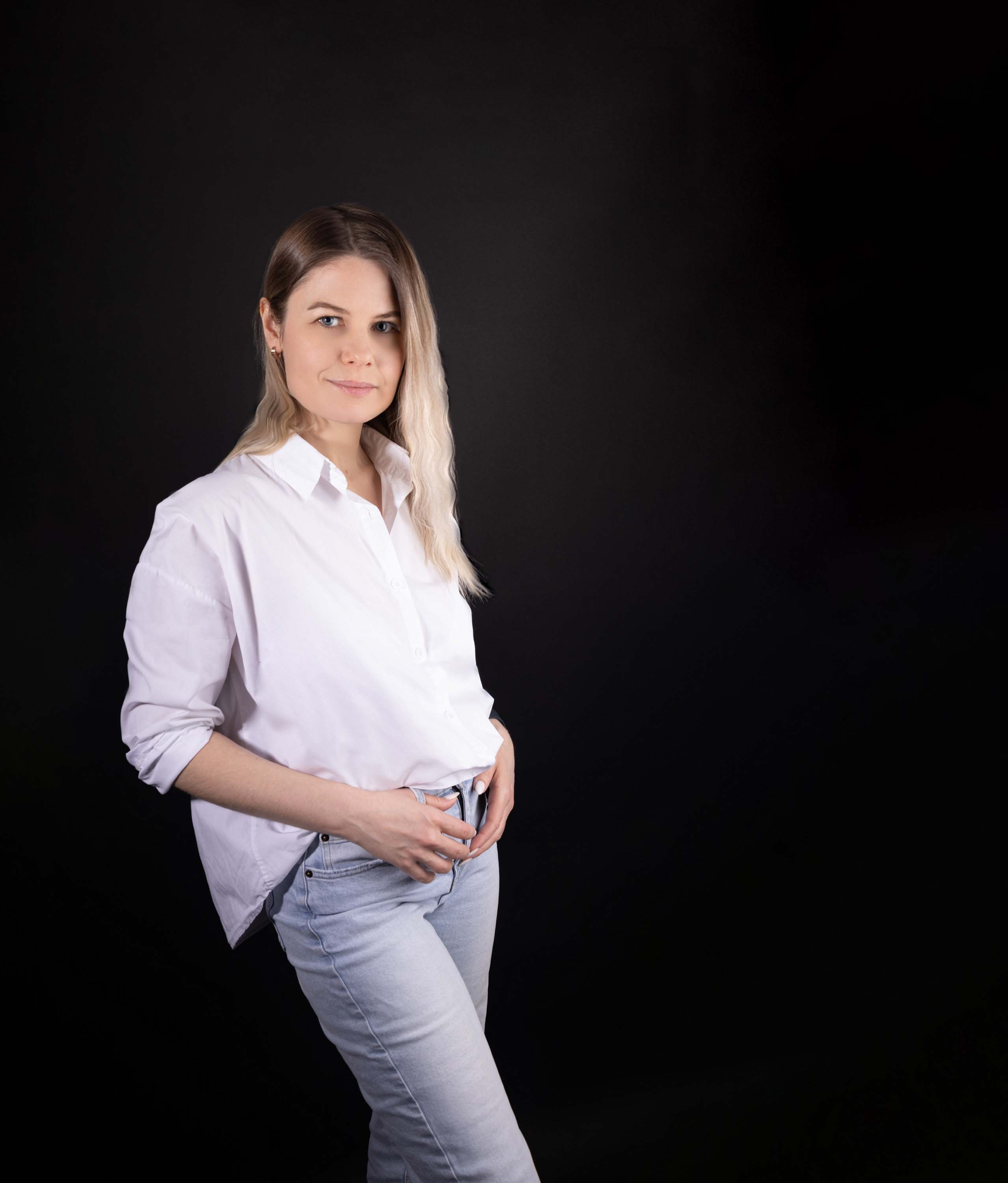 Светлые волосы, белая рубашка, джинсы, уверенная поза, тёмный фон.