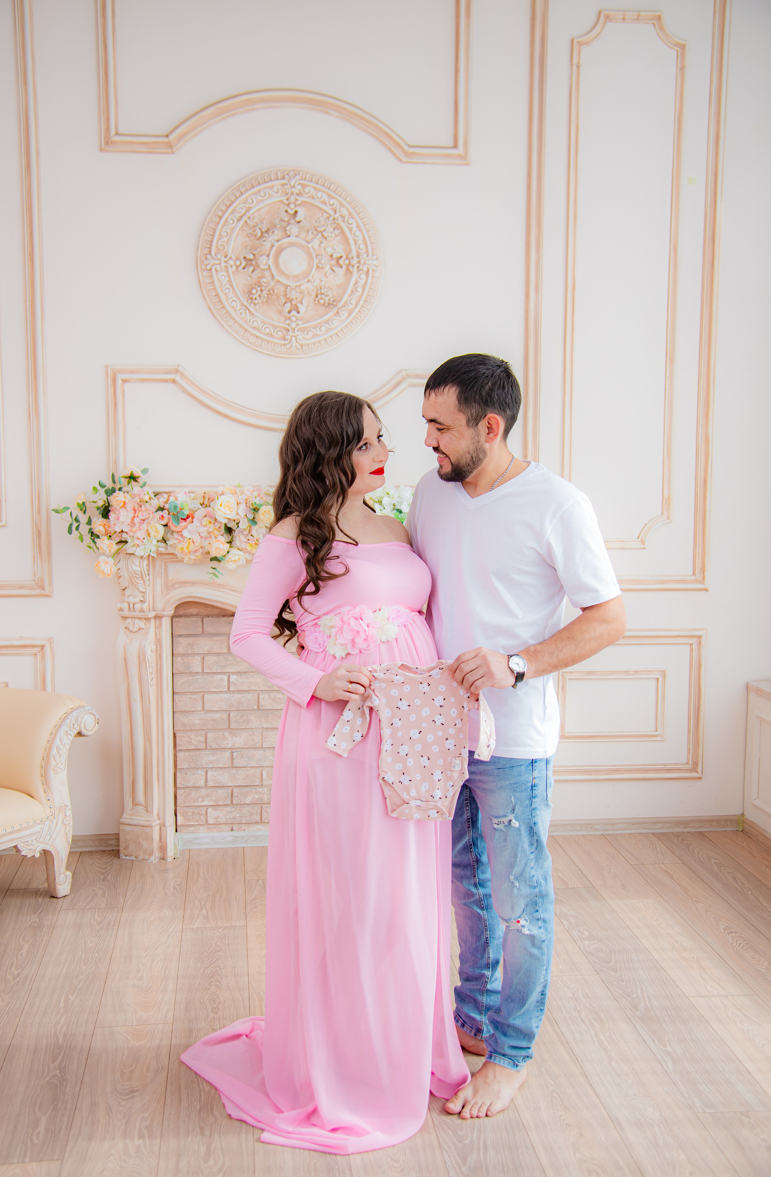 Фотосессия беременных в фотостудии в Уфе: будущая мама в элегантном платье из проката, с профессиональным макияжем и прической. Она счастливо позирует на фоне стильного интерьера, а рядом ее муж и члены семьи. Фотограф помогает в позировании, создавая теплую и уютную атмосферу, отражающую красоту и нежность материнства