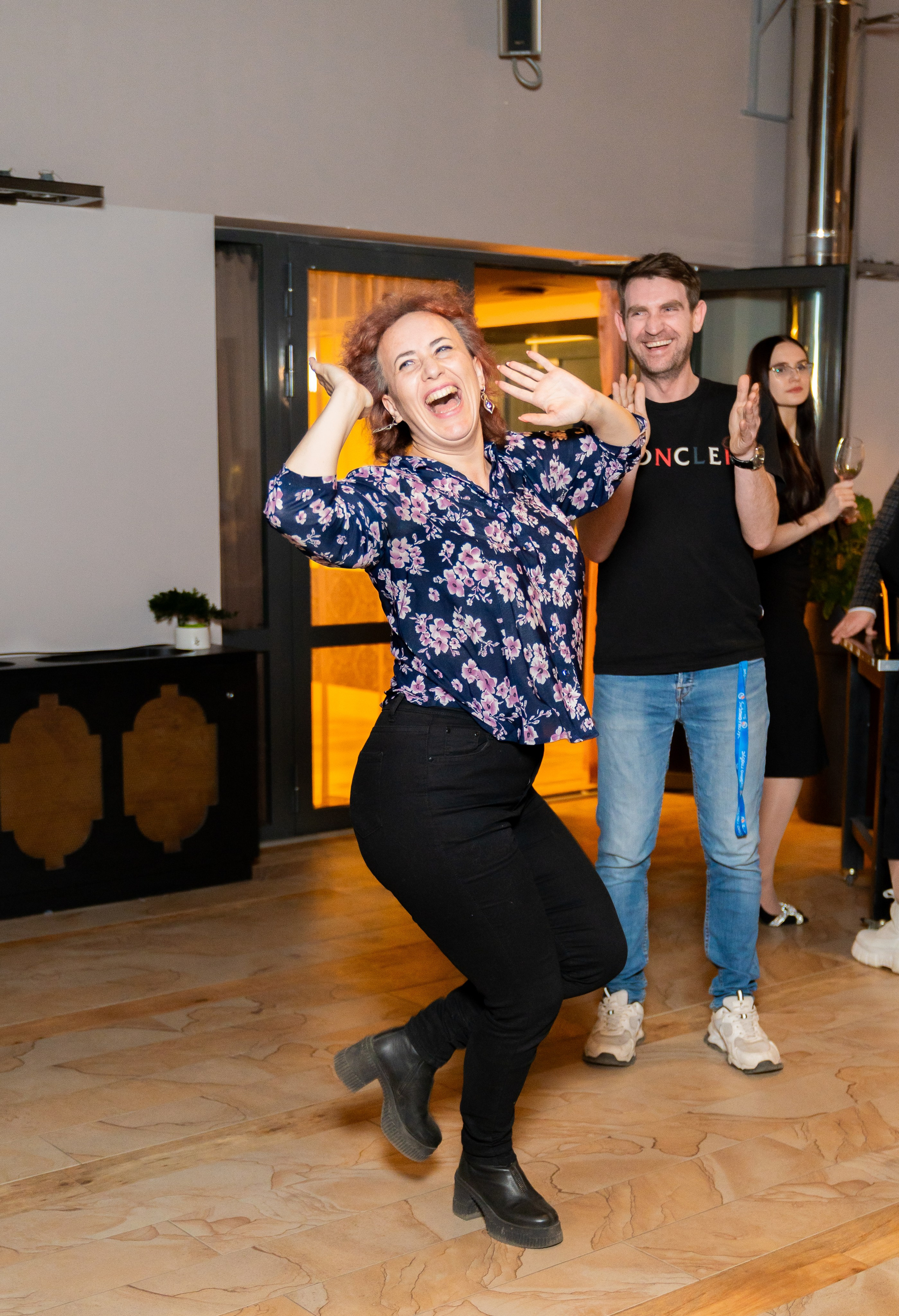 Эмоциональная гостья в синей цветочной блузке зажигательно танцует на вечеринке в Radisson Sonya. Рядом улыбается мужчина в футболке Moncler. Фото Евгения Мальцева, evrednix.ru.