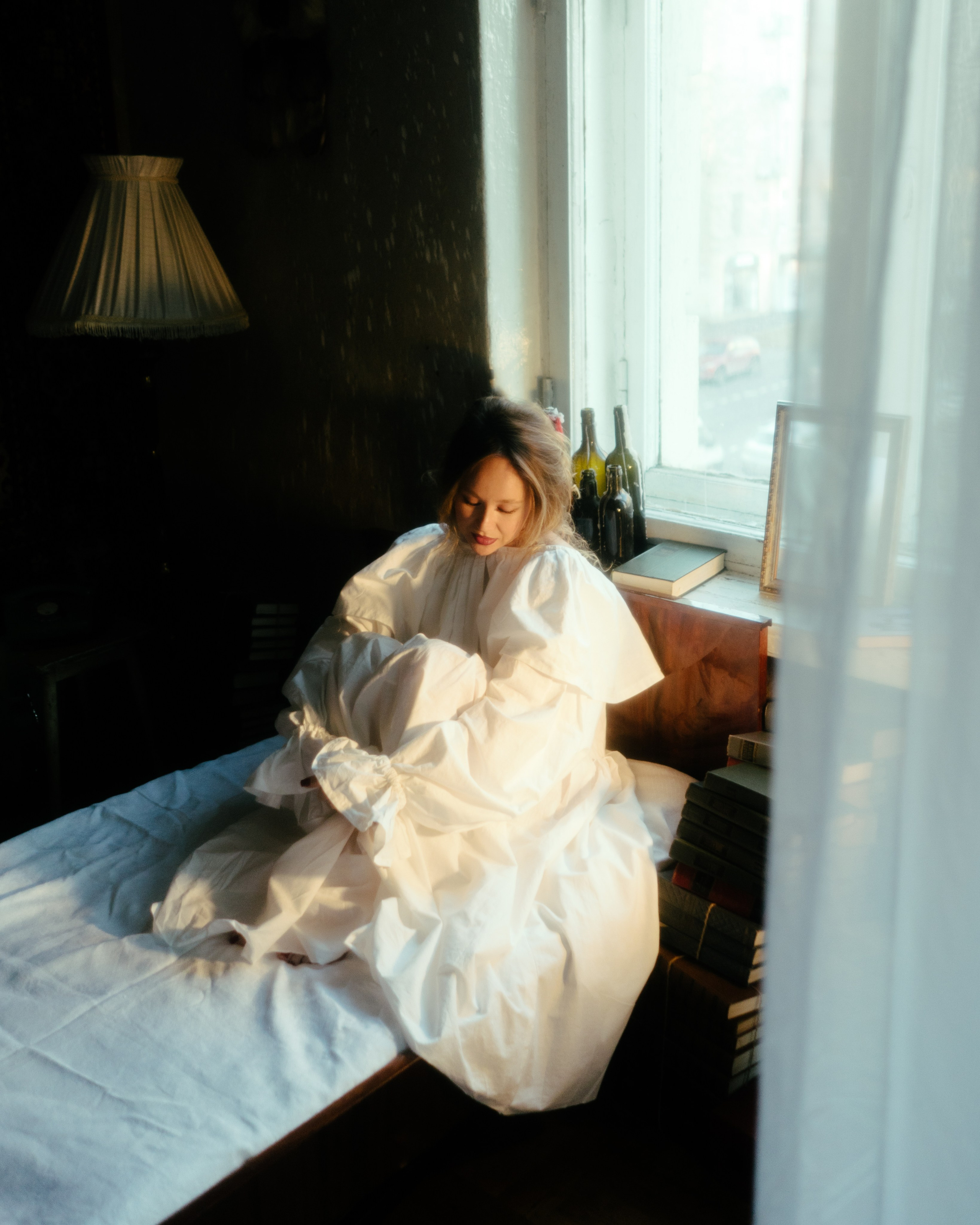 Нежный портрет беременности на кровати: модель в воздушном винтажном белом платье у окна, мягкий свет, уютная студийная съемка в Санкт-Петербурге, кадры как в кино.