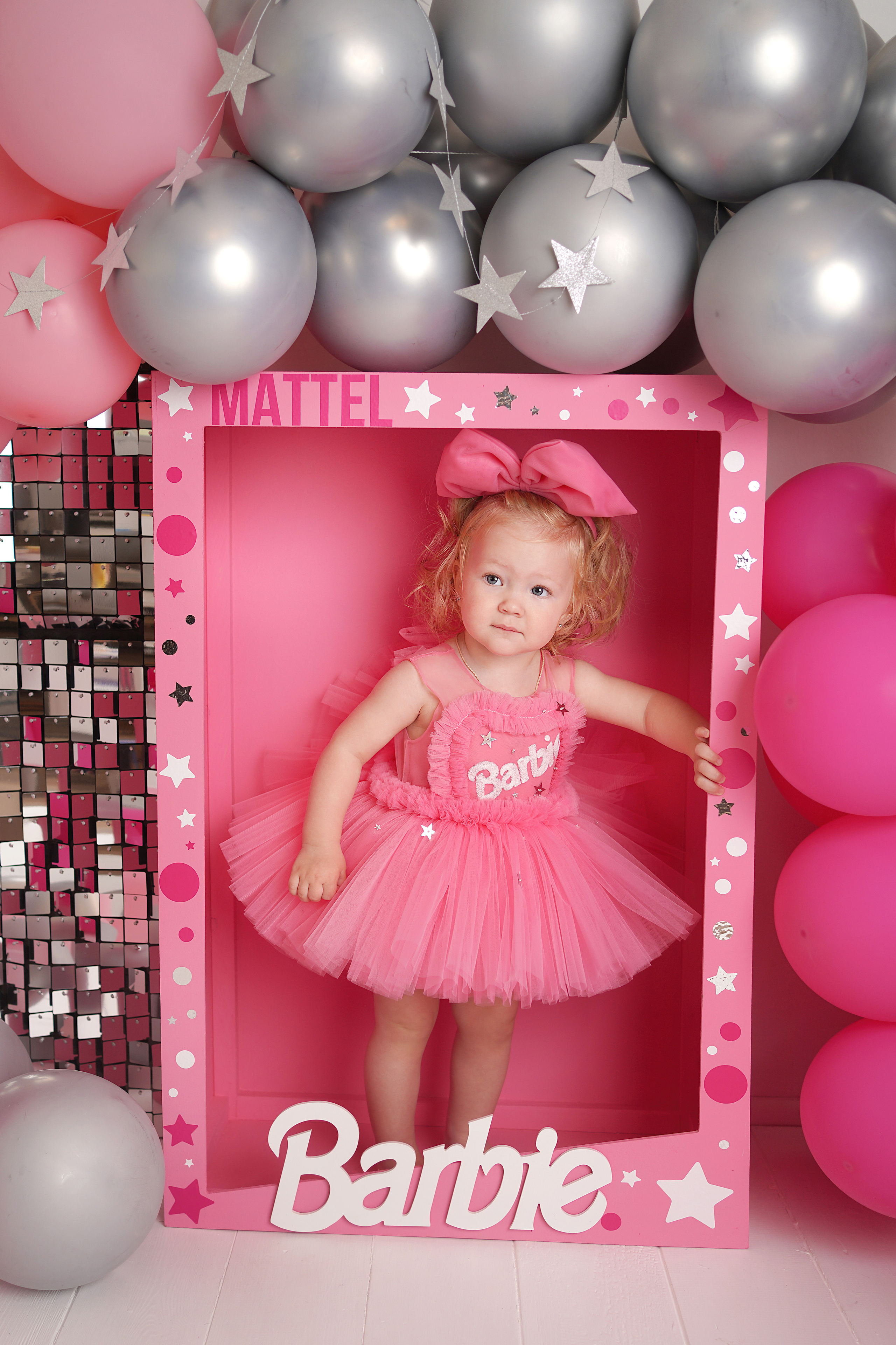 Малышка стоит в  большой коробке Барби. На девочке розовое платье и бант.