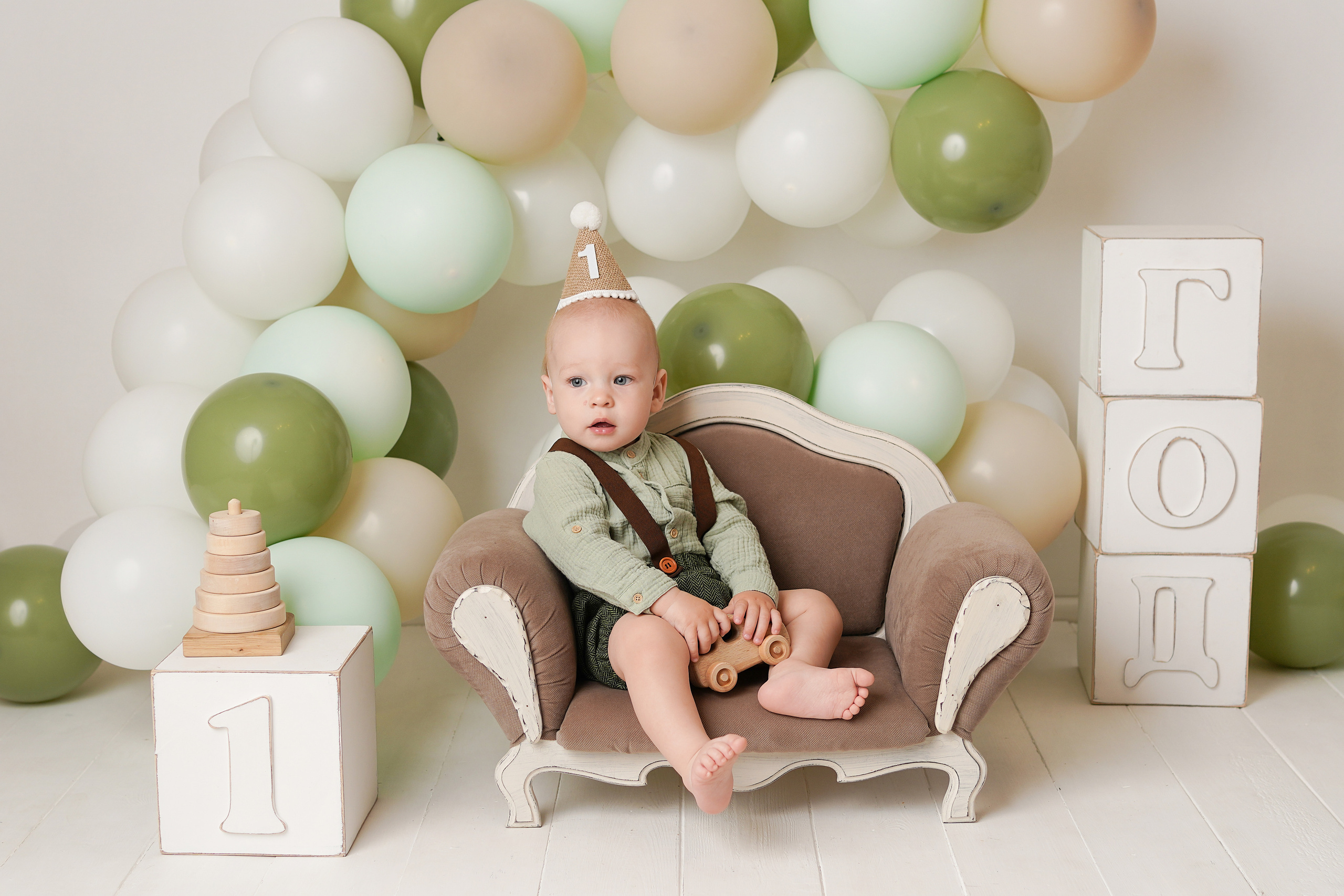 На фото мальчик сидит на крошечном детском диванчике, рядом много шариков, огоньки и кубики год.