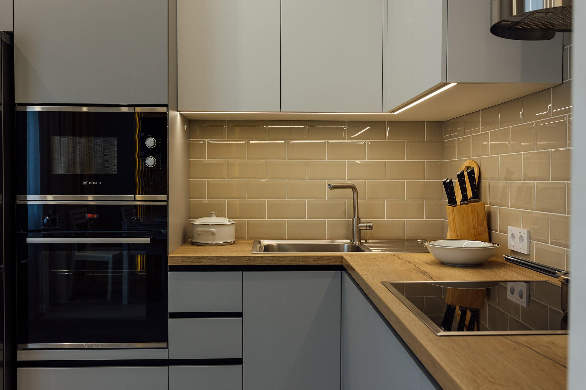 Фотография угловой кухни в современном стиле: серые матовые фасады без ручек в сочетании со столешницей под дерево. Рабочая зона с подсветкой и фартуком из глянцевой плитки. Фото для каталога кухонной мебели в Пензе.