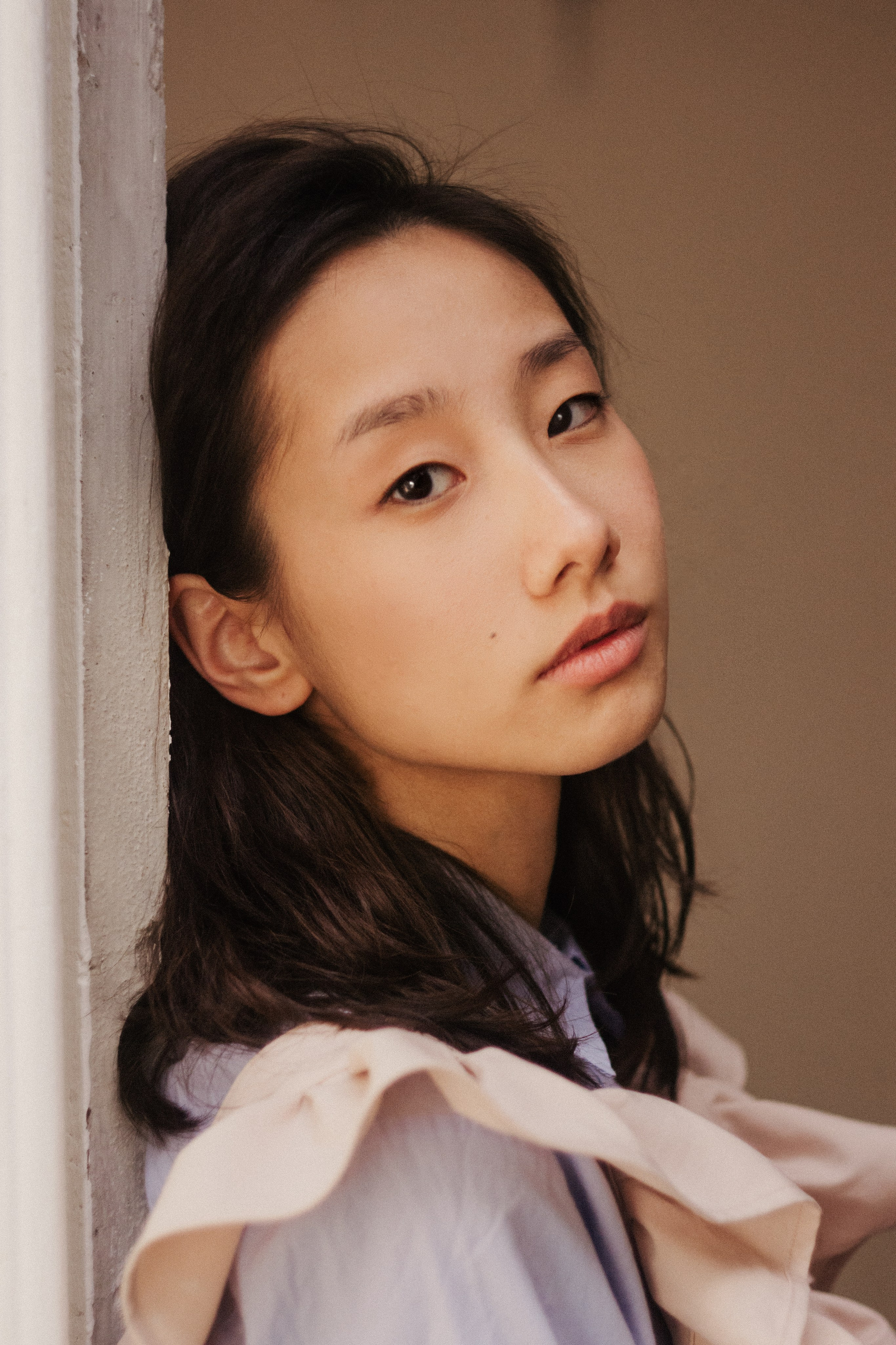 Крупный портрет девушки, которая опирается на косяк двери и смотри на нас. У нее темные волосы, светло-голубая рубашка и объемный светло-розовый передник