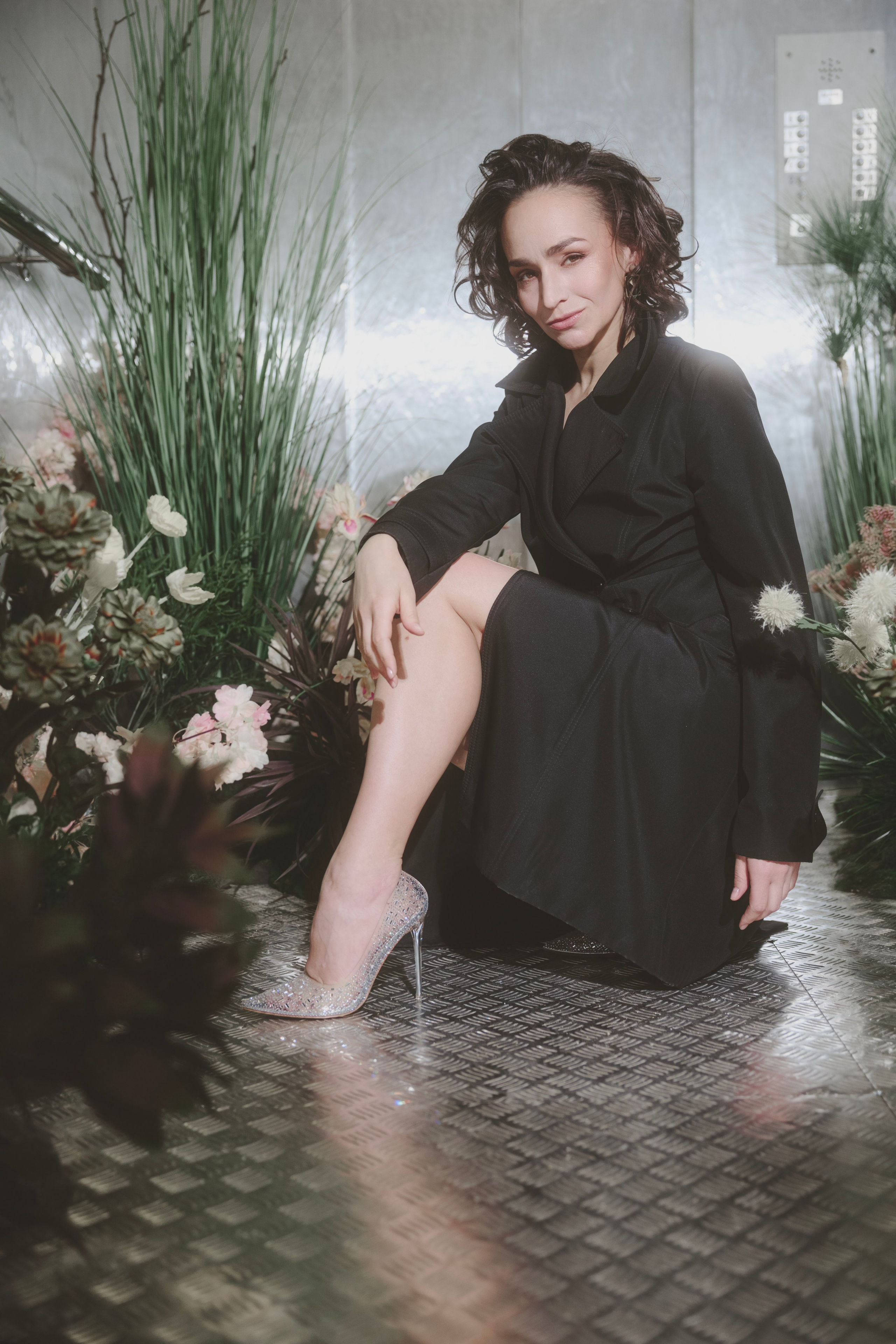 Портретное фото молодой девушки в студии. Девушка сидит на фоне нейтрального цвета, ее лицо освещено мягким светом. Она смотрит прямо в камеру с легкой улыбкой. На девушке стильная одежда, подчеркивающая современный модный стиль. Фотография выполнена в высоком разрешении, с акцентом на деталях