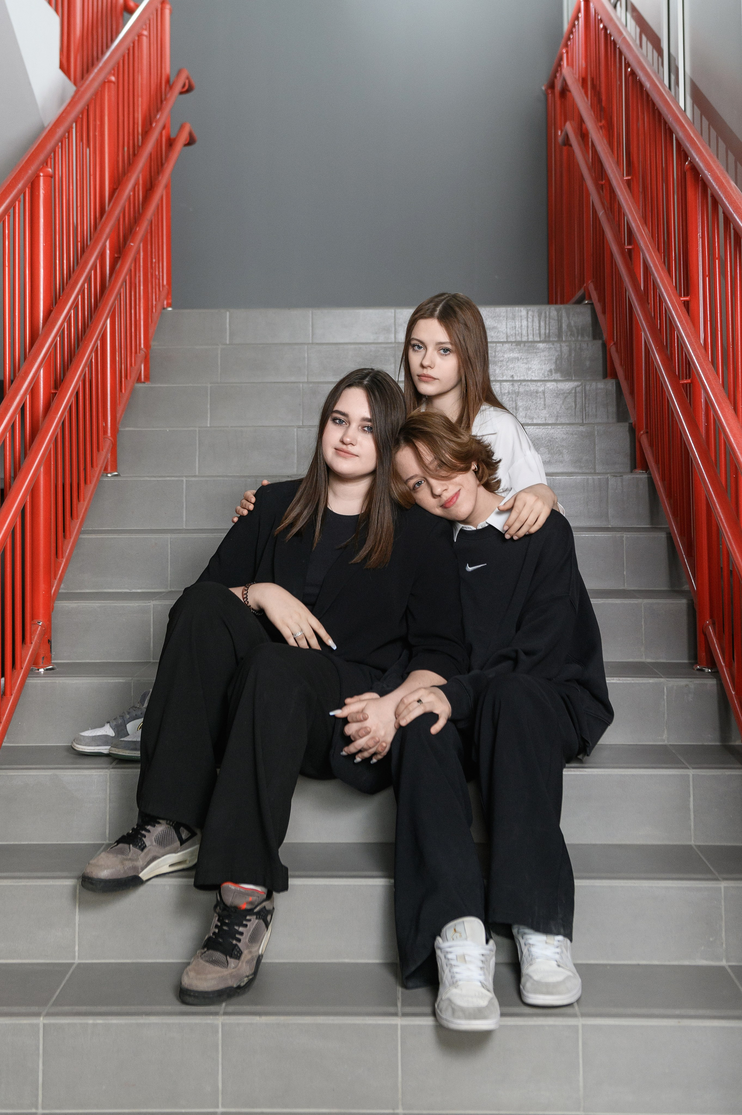 Три девушки сидят на серой лестнице с ярко-красными перилами.