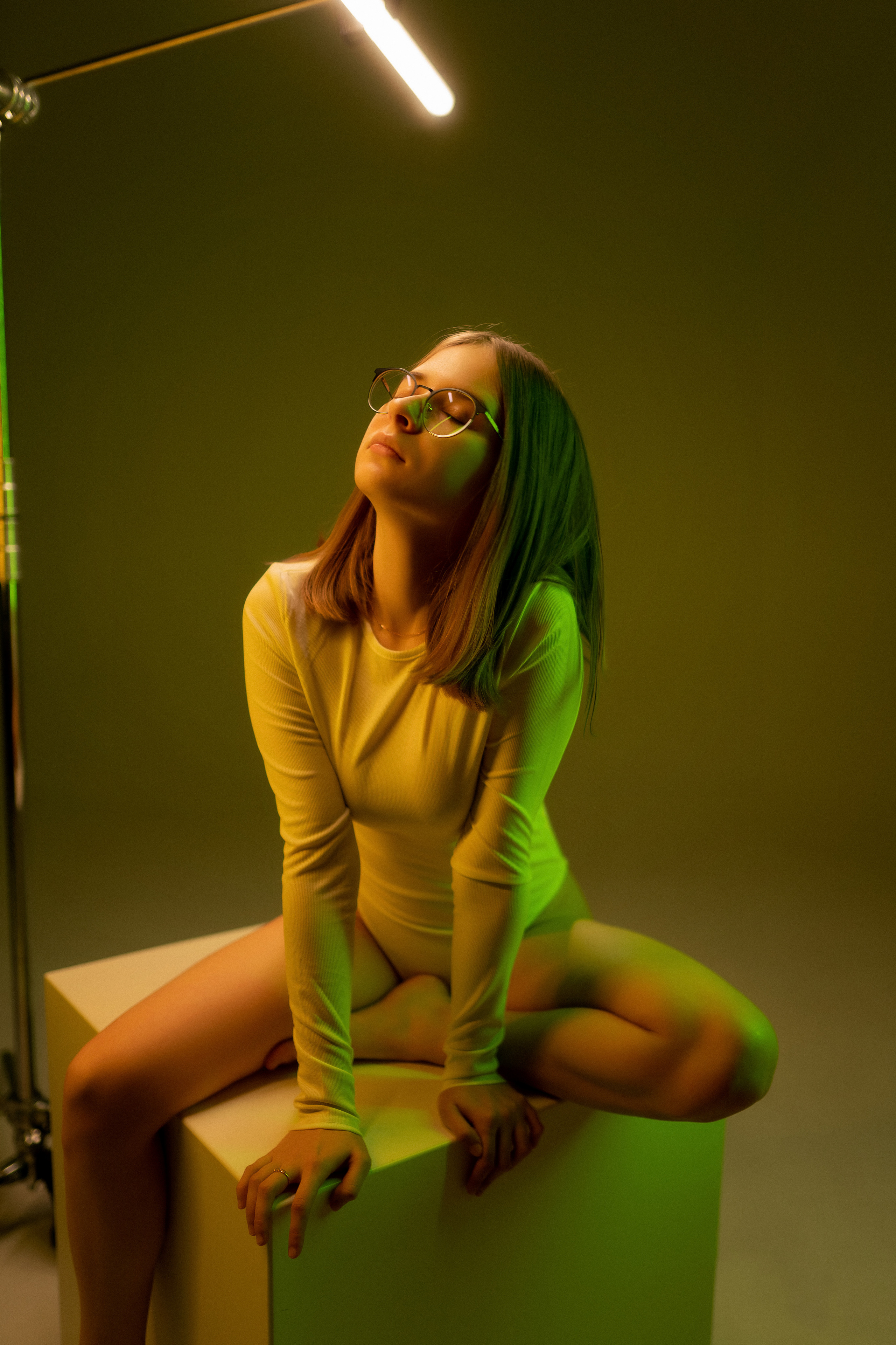 женский ростовой портрет красивая девушка в белом боди сидит на кубе в студии с циклорамой освещена цветным светом