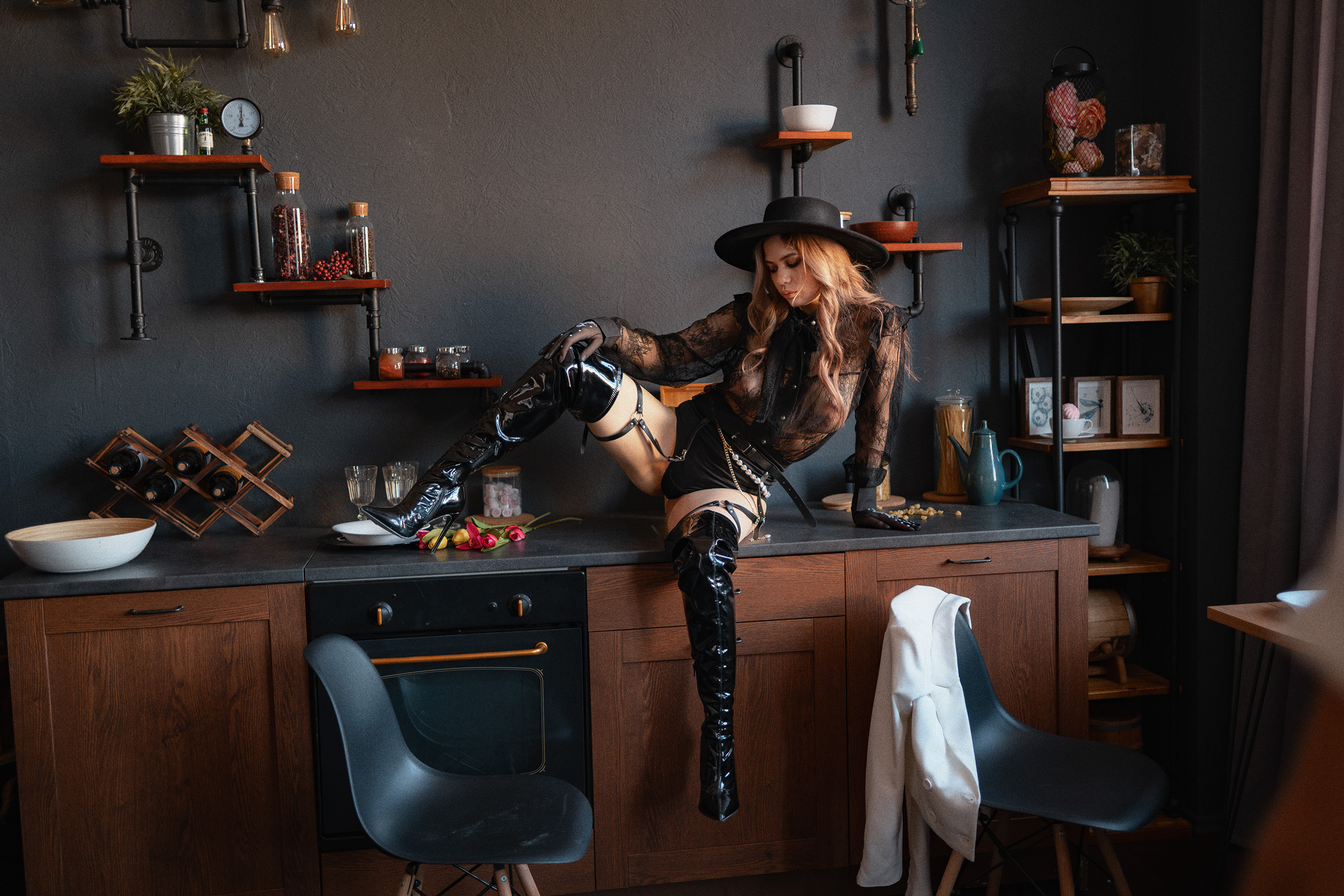 Девушка в черном эротическом наряде и черных сапогах и шляпе сидит на кухонном гарнитуре