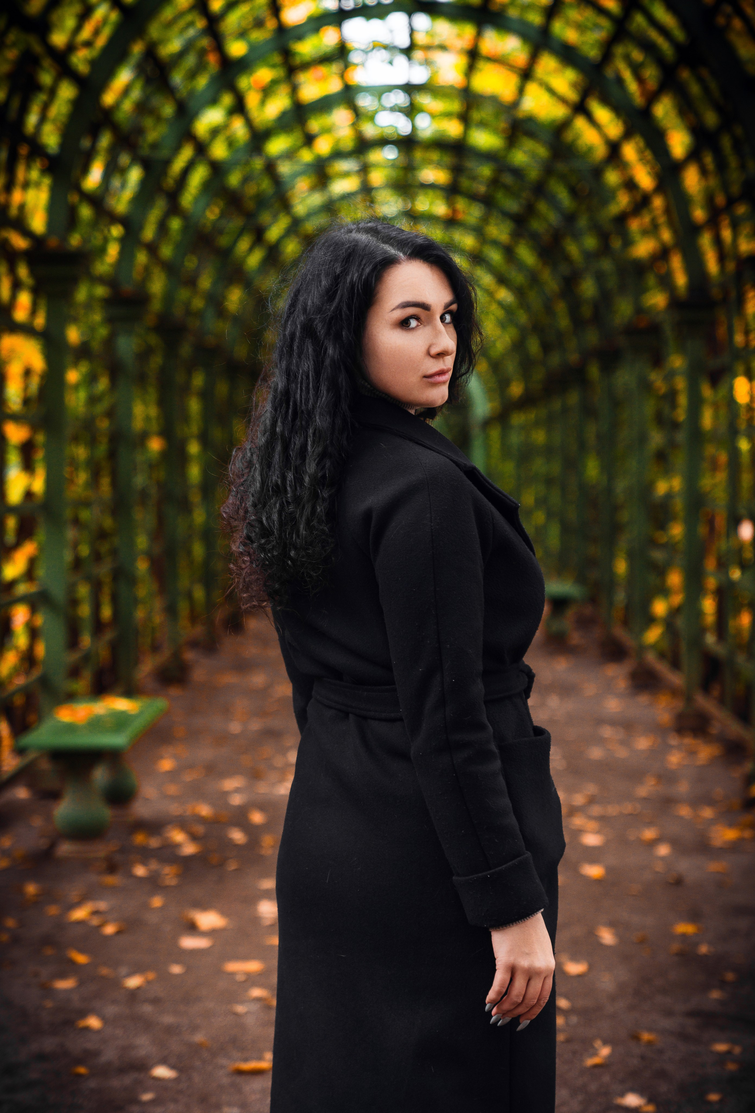 Осенний Санкт-Петербург. Летний Сад. Элегантная брюнетка в чёрном пальто позирует под зелёной аркой в осеннем парке, окружённая жёлтыми и оранжевыми листьями. Фотограф Мальцев Евгений.