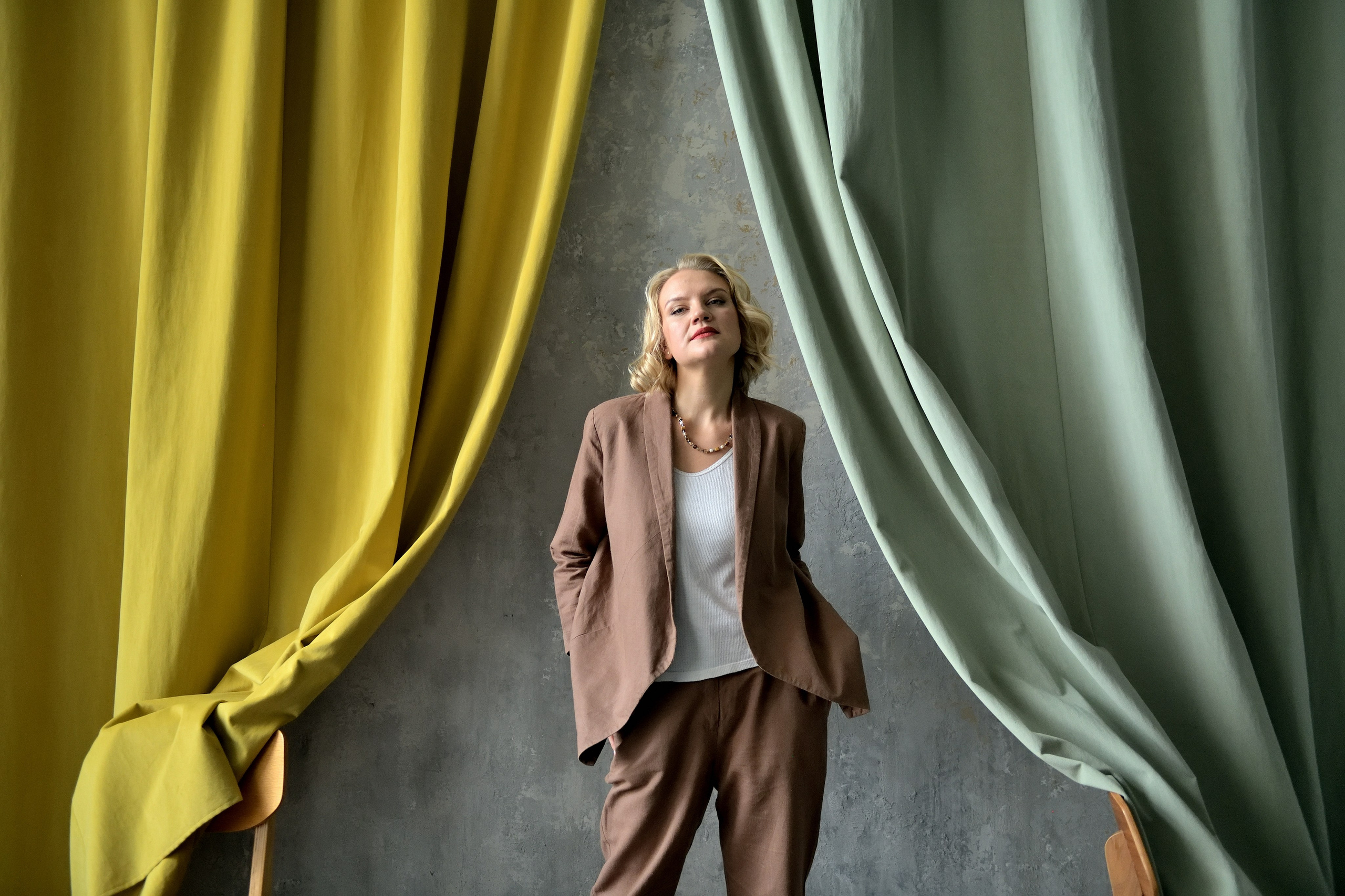 Девушка в костюме цвета Pantone 2025 стоит на фоне зелено-желтой шторы.