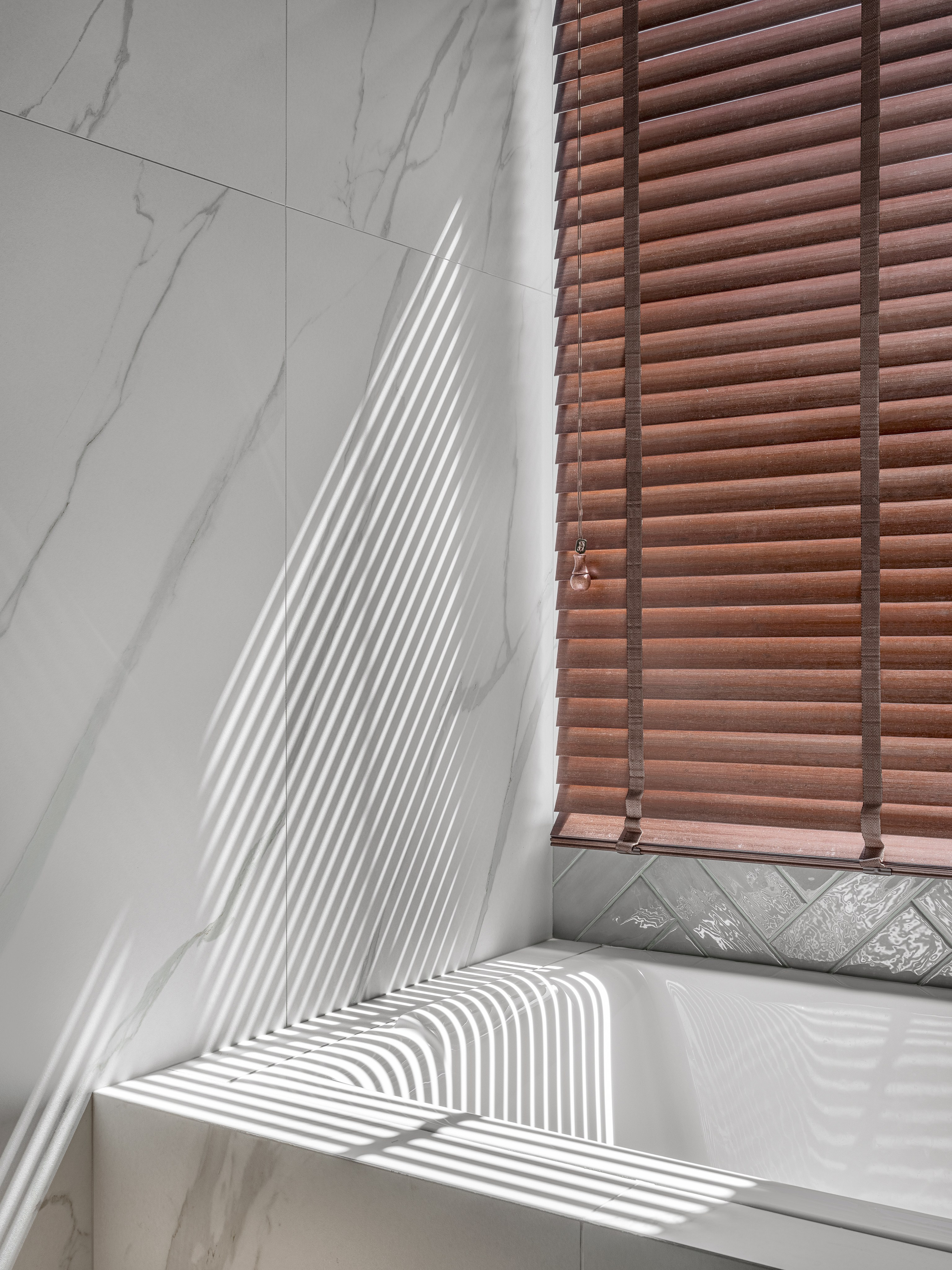 Утончённый интерьер ванной комнаты с белой плиткой под мрамор, ванной и деревянными жалюзи, создающими красивую игру света и тени, дизайнеры Алёна Попова и Анастасия Дмитриева, фотограф Сергей Денисенко, современный стиль.