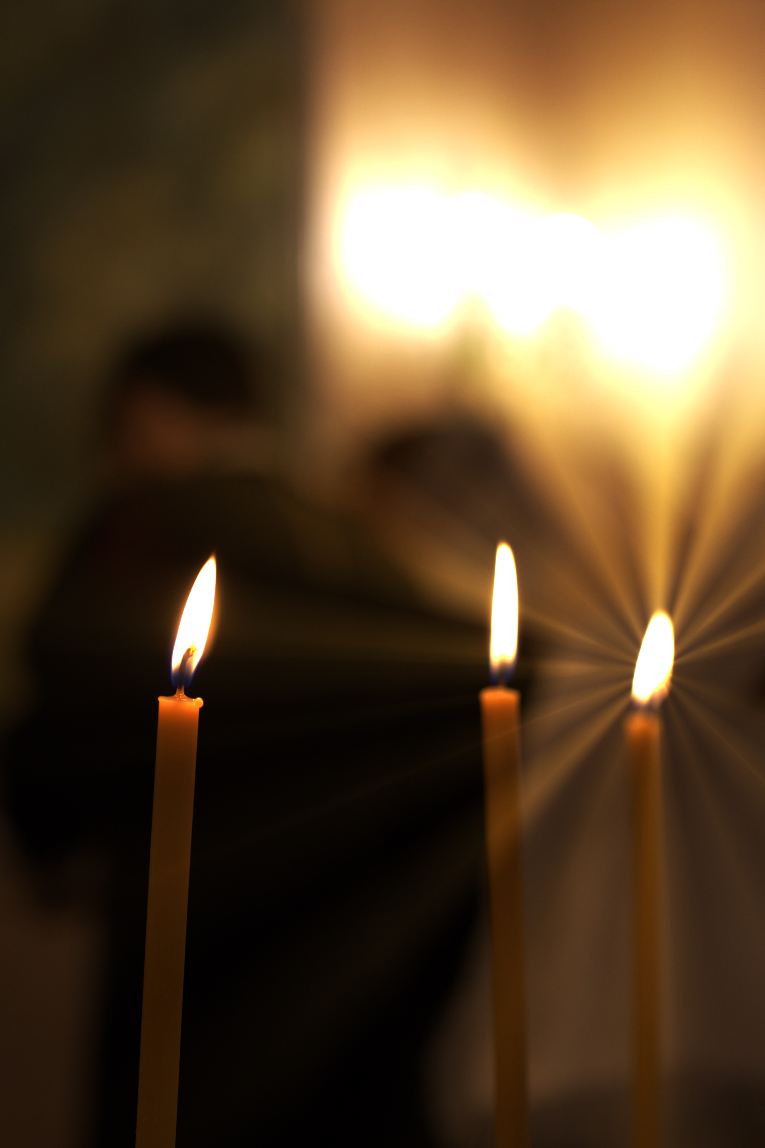 Три горящие церковные свечи на переднем плане, на заднем фоне размытые фигуры людей во время таинства крещения ребенка
