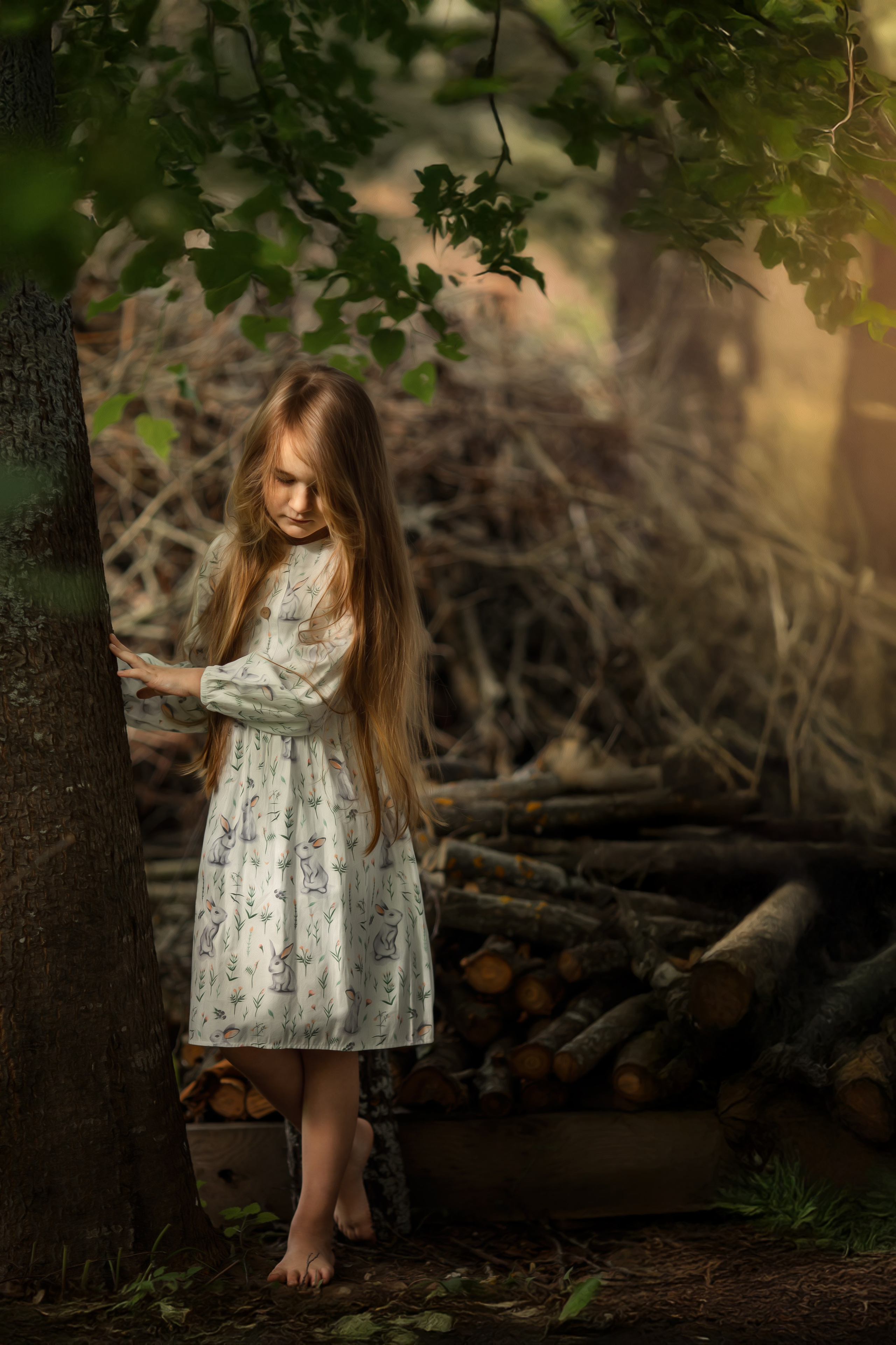 Девочка с длинными светлыми волосами в платье с зайцами стоит у дерева на фоне сложенных веток. Мягкий свет пробивается сквозь листву, создавая сказочную лесную атмосферу.