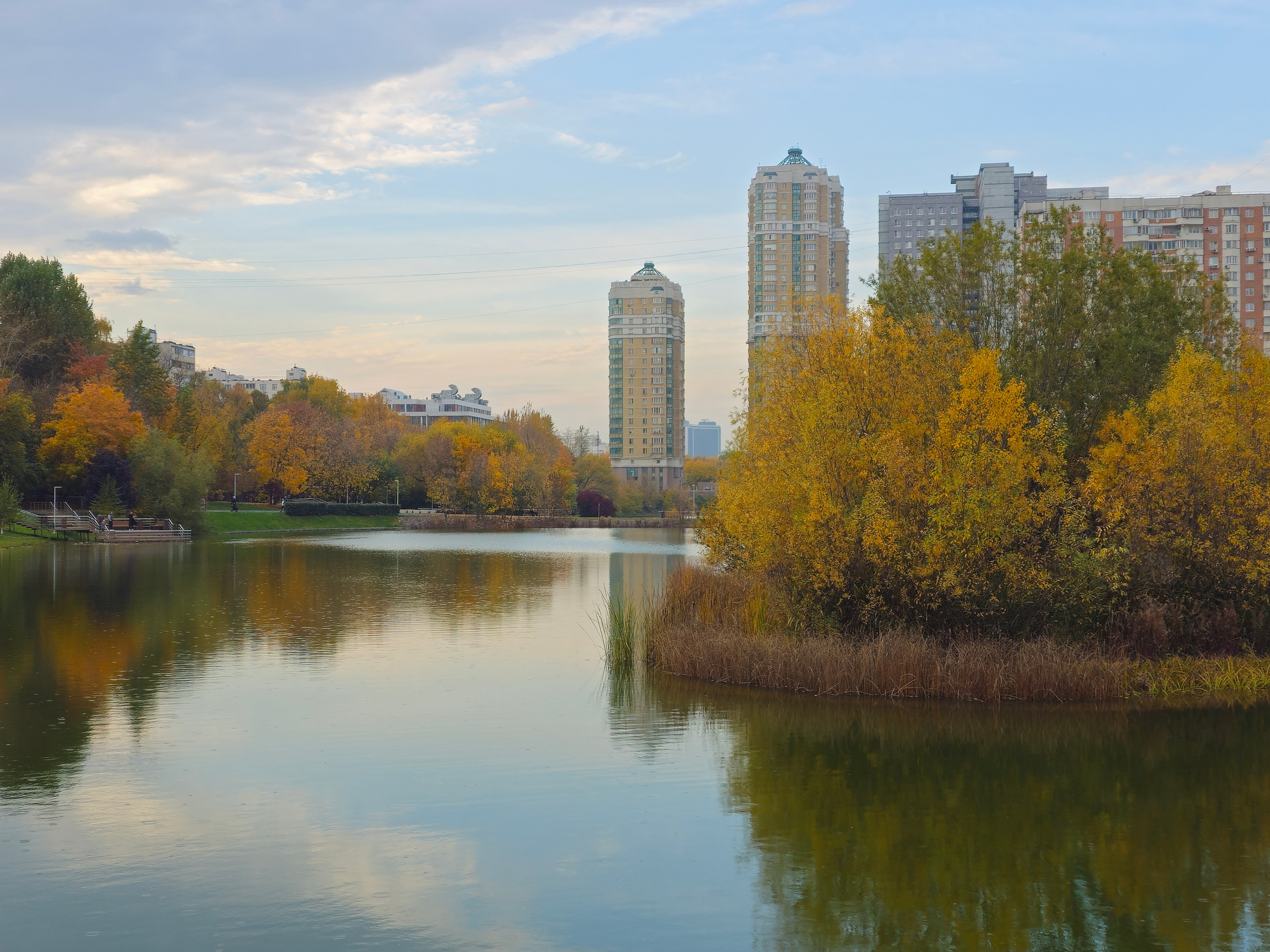 Пейзаж городского водоема с отражением в воде. На дальнем берегу расположена линия деревьев с желтой и красной листвой, а за ними возвышаются современные высотные жилые дома, включая два заметных здания с бирюзовыми крышами.