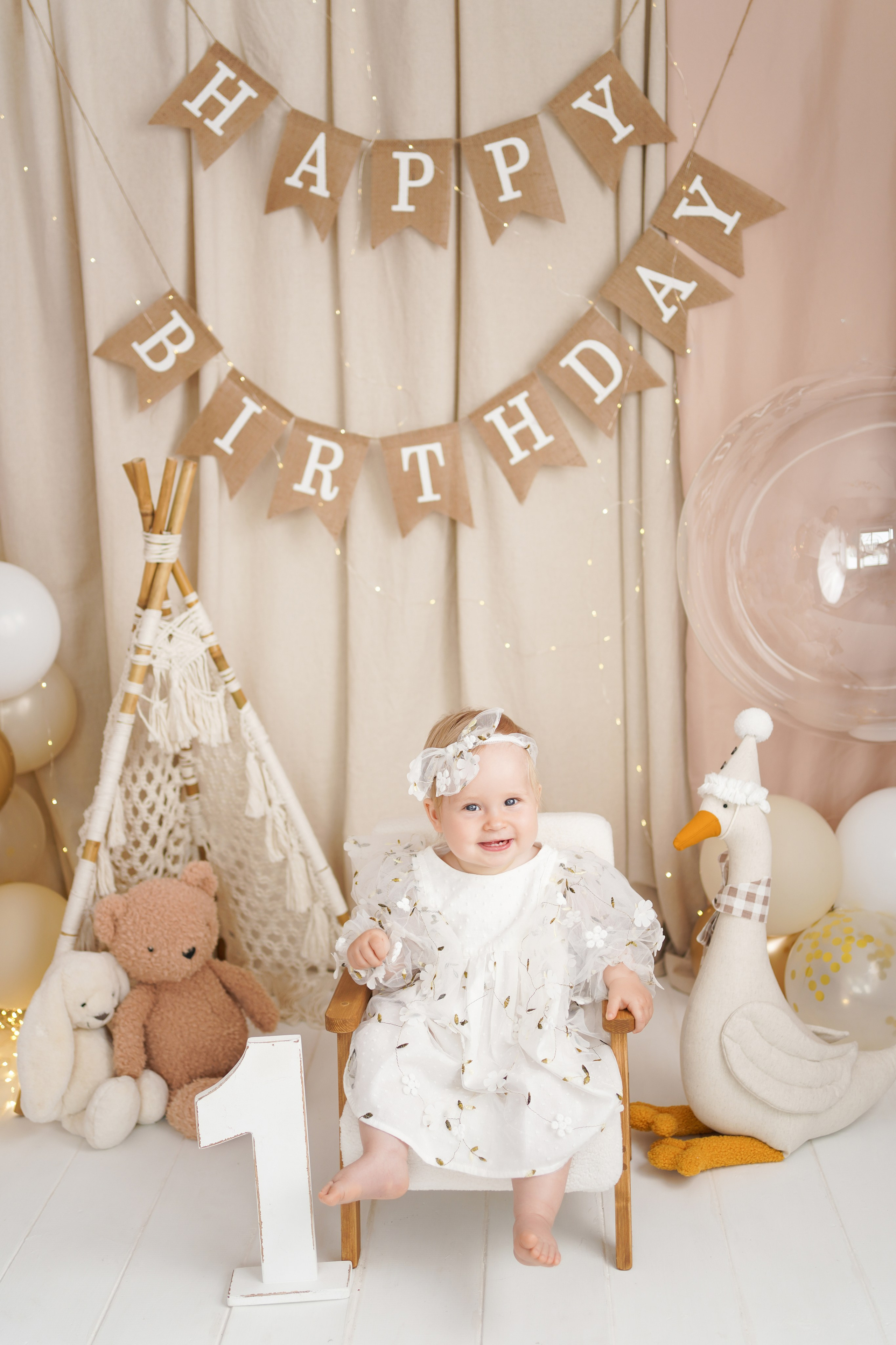 На фото улыбающаяся малышка в нарядной одежде. Она сидит на детском стульчике. Рядом гусь, мишка, зайка.