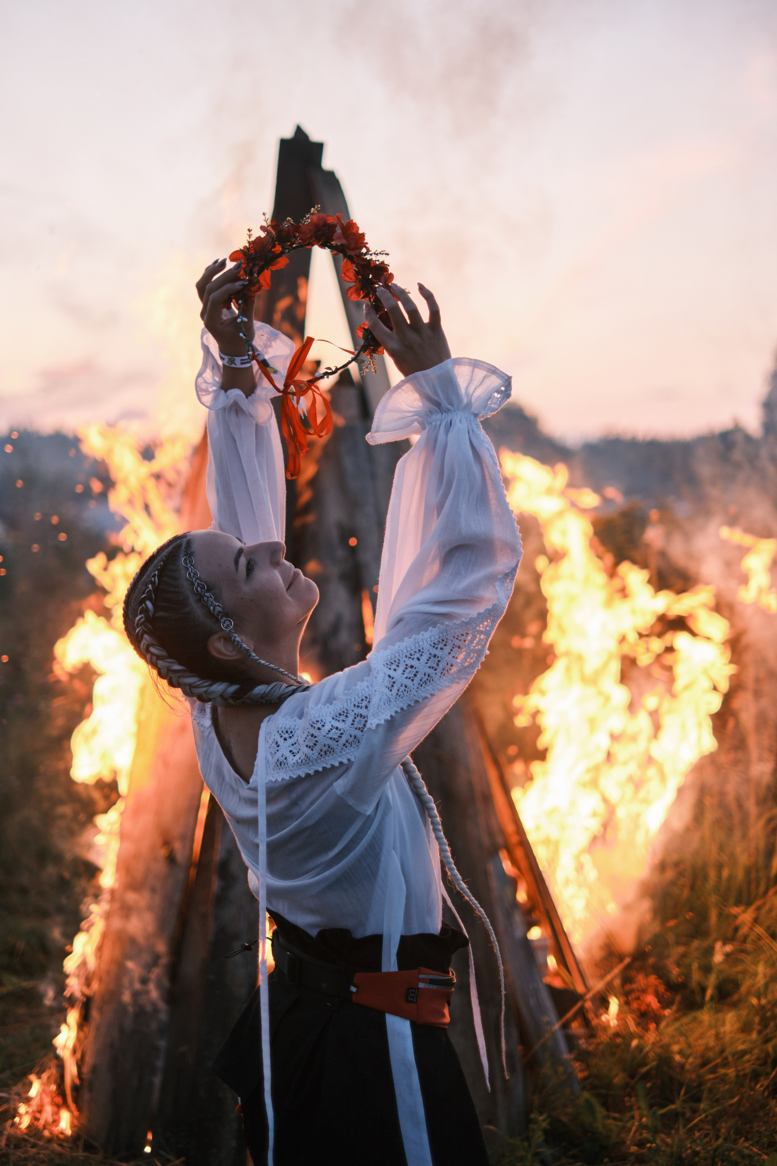 Девушка стоит в русском костюме и венком сзади горит большой костер
