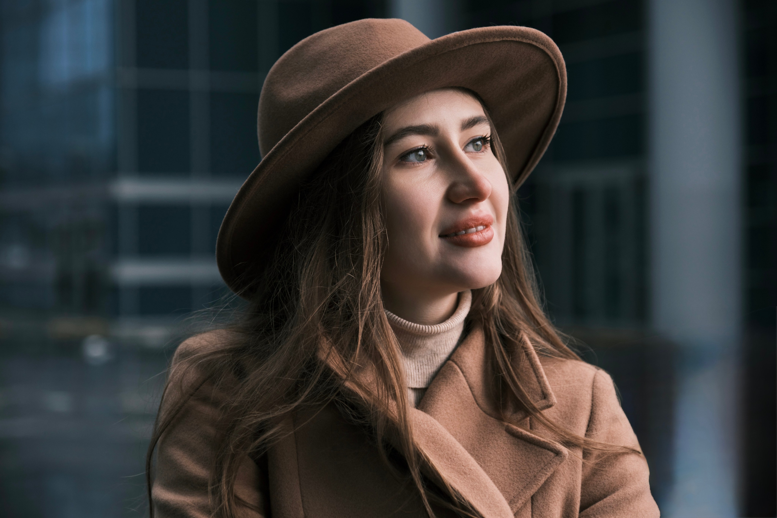 Крупный план: портрет девушки в коричневой шляпе, смотрящей в сторону, на размытом фоне городских зданий.