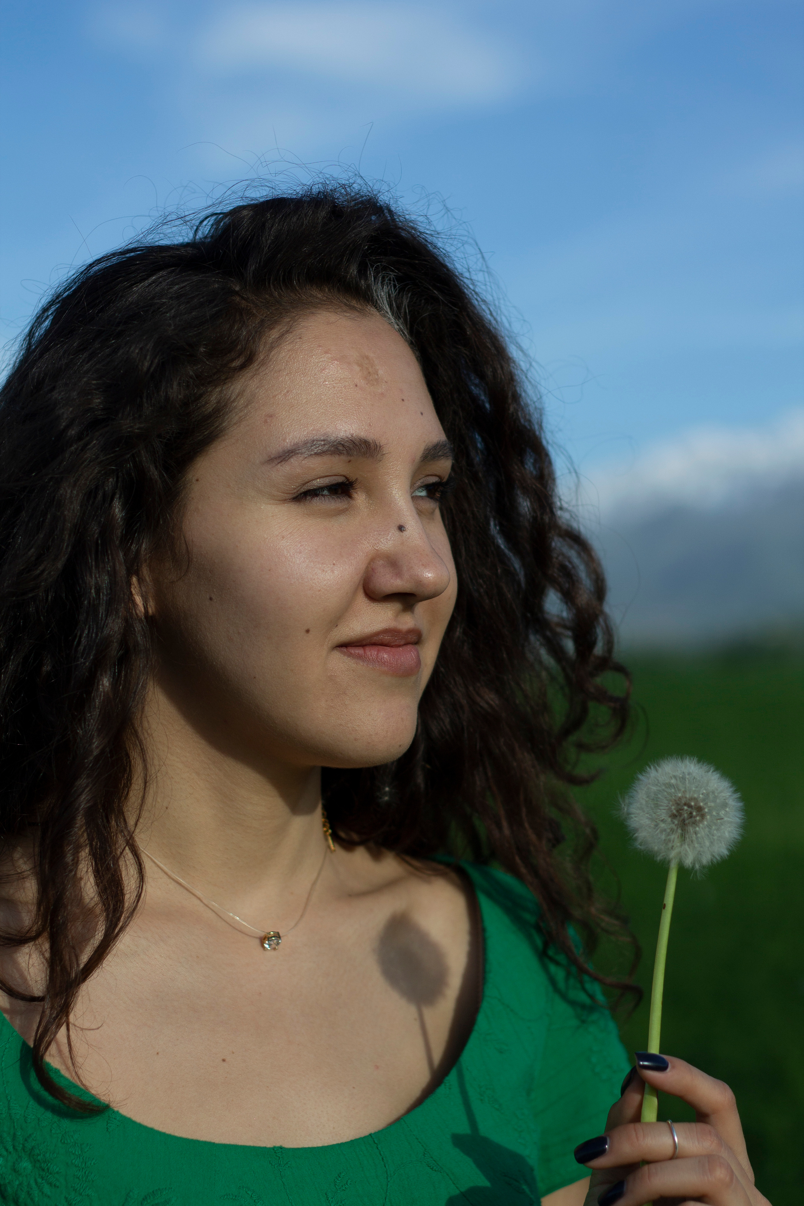 Длинноволосая кудрявая девушка в зеленом платье смотрит на весенний одуванчик