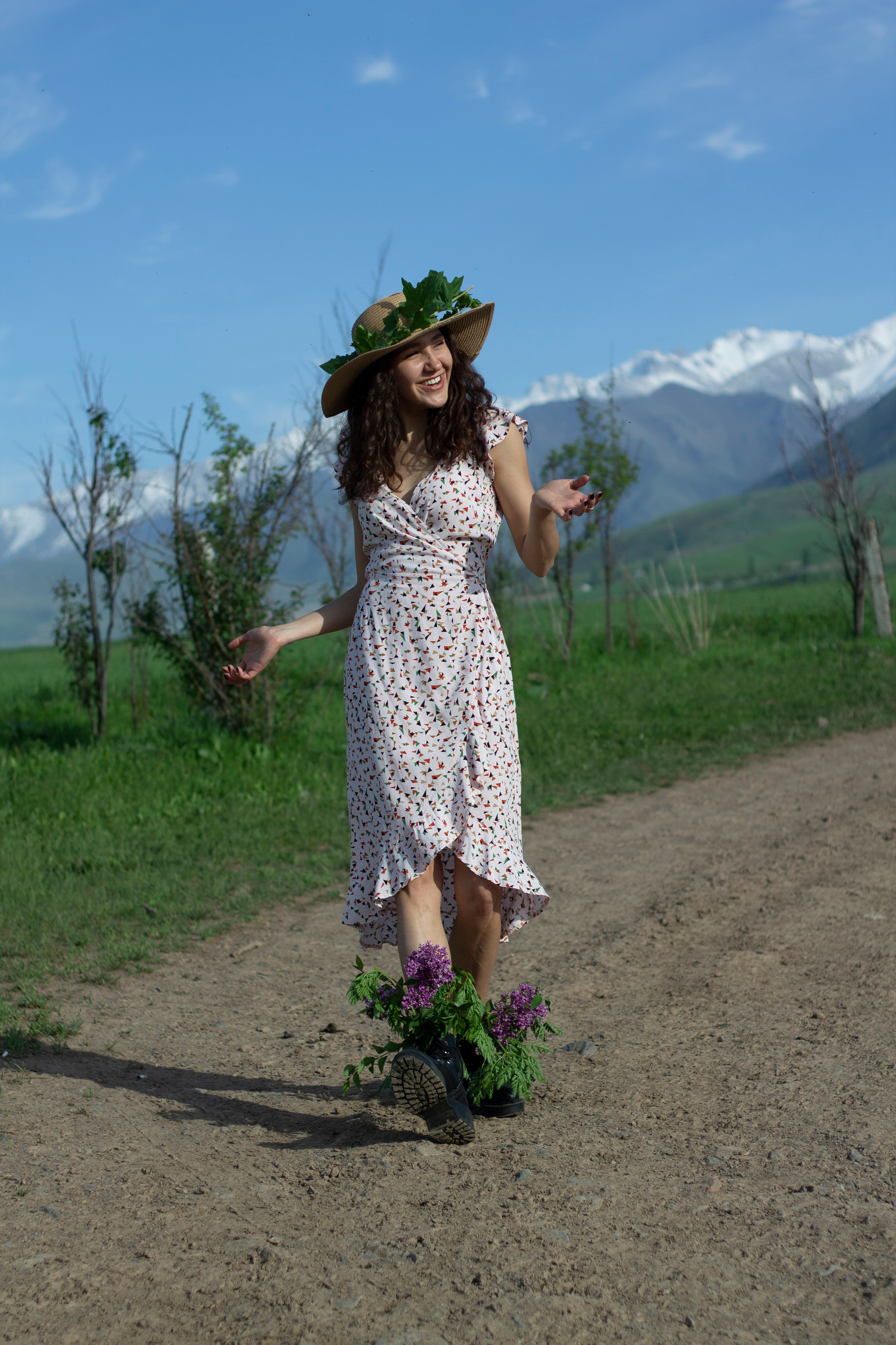 Девушка в платье в цветочек несет цветы в ботинках и венок на шляпе 