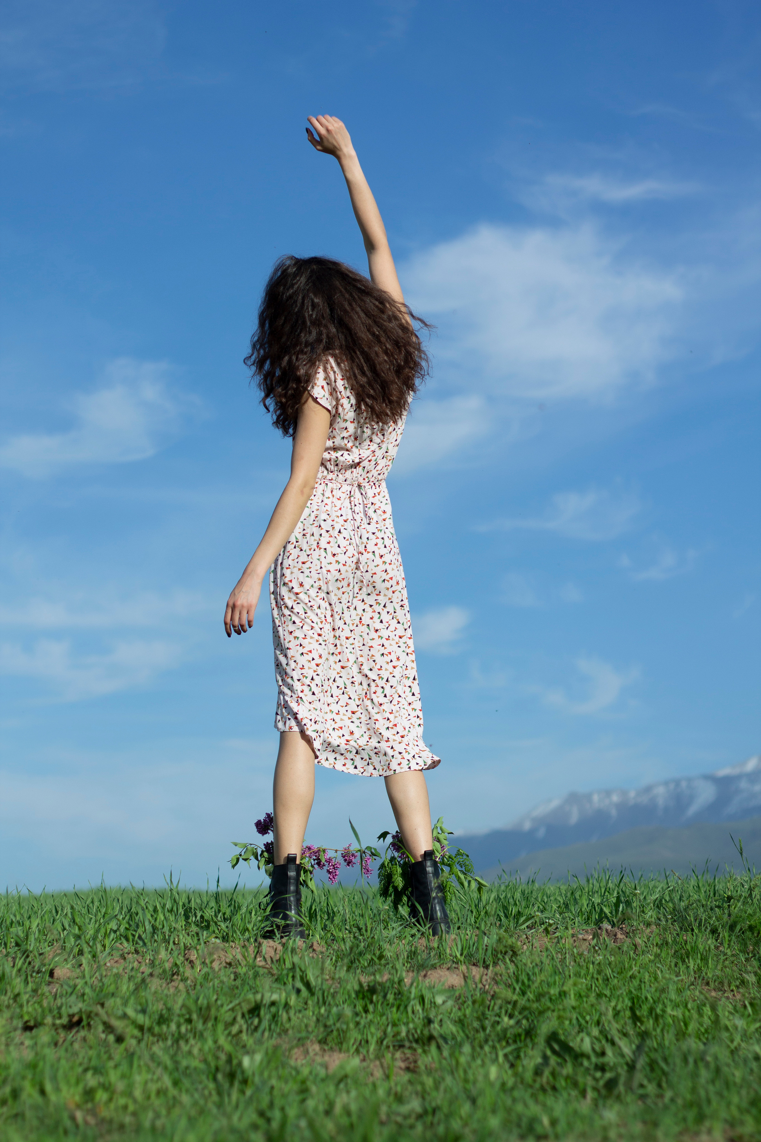 Девушка в платье в цветочек стоит спиной к фотографу и поднимает правую руку вверх