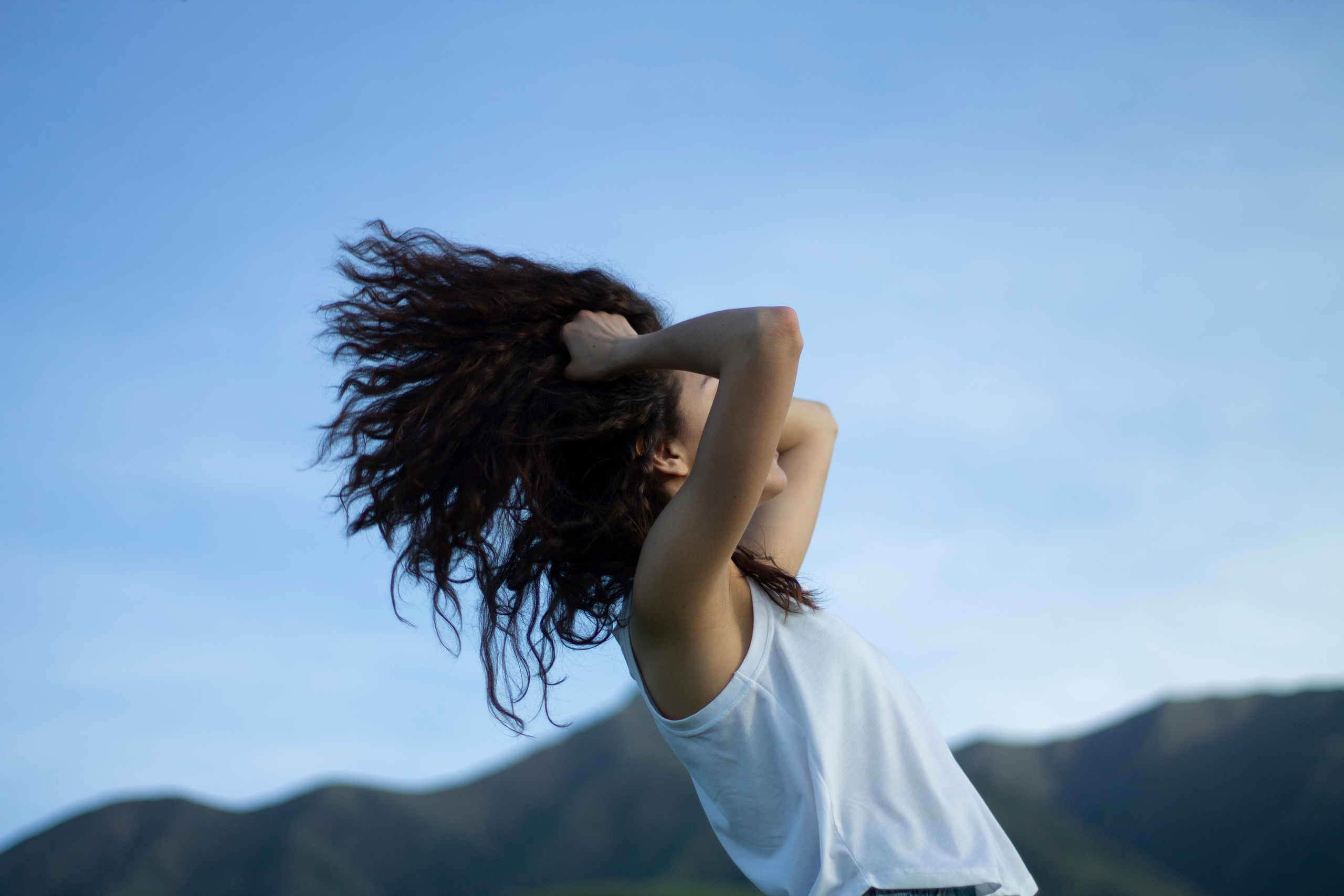 На фоне холмов Кыргызстана девушка в белой майке на фоне гор машет волосами и держит руки у головы