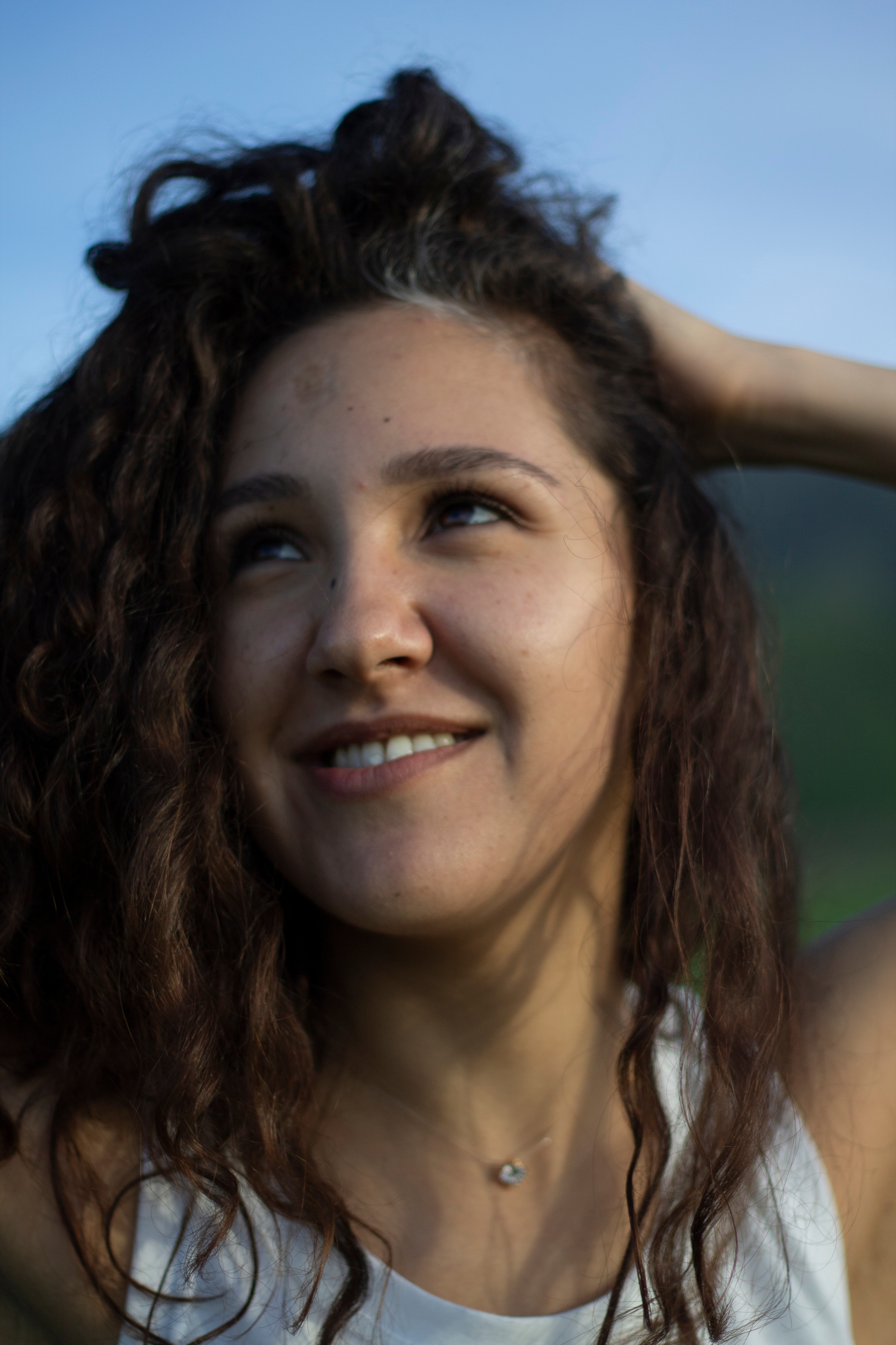 Девушка в белой майке мечтательно смотрит в небо и держит волосы рукой