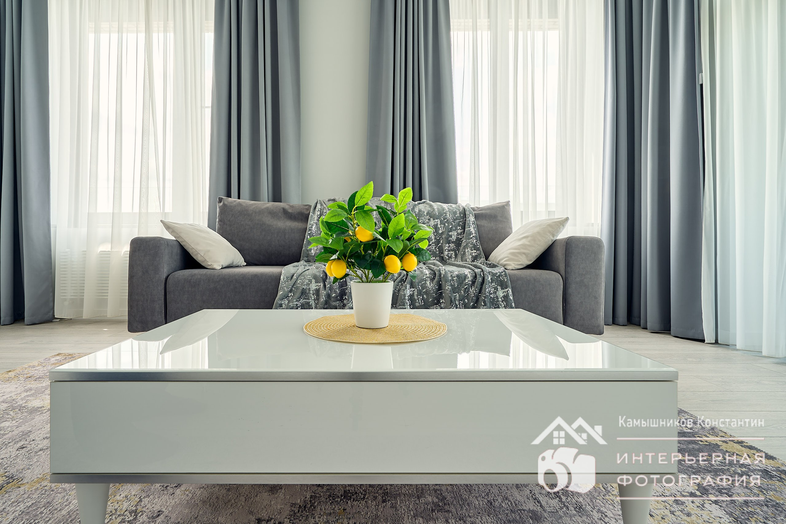 Интерьерная съемка с крупным планом журнального столика , за ним - дивана и двух окон