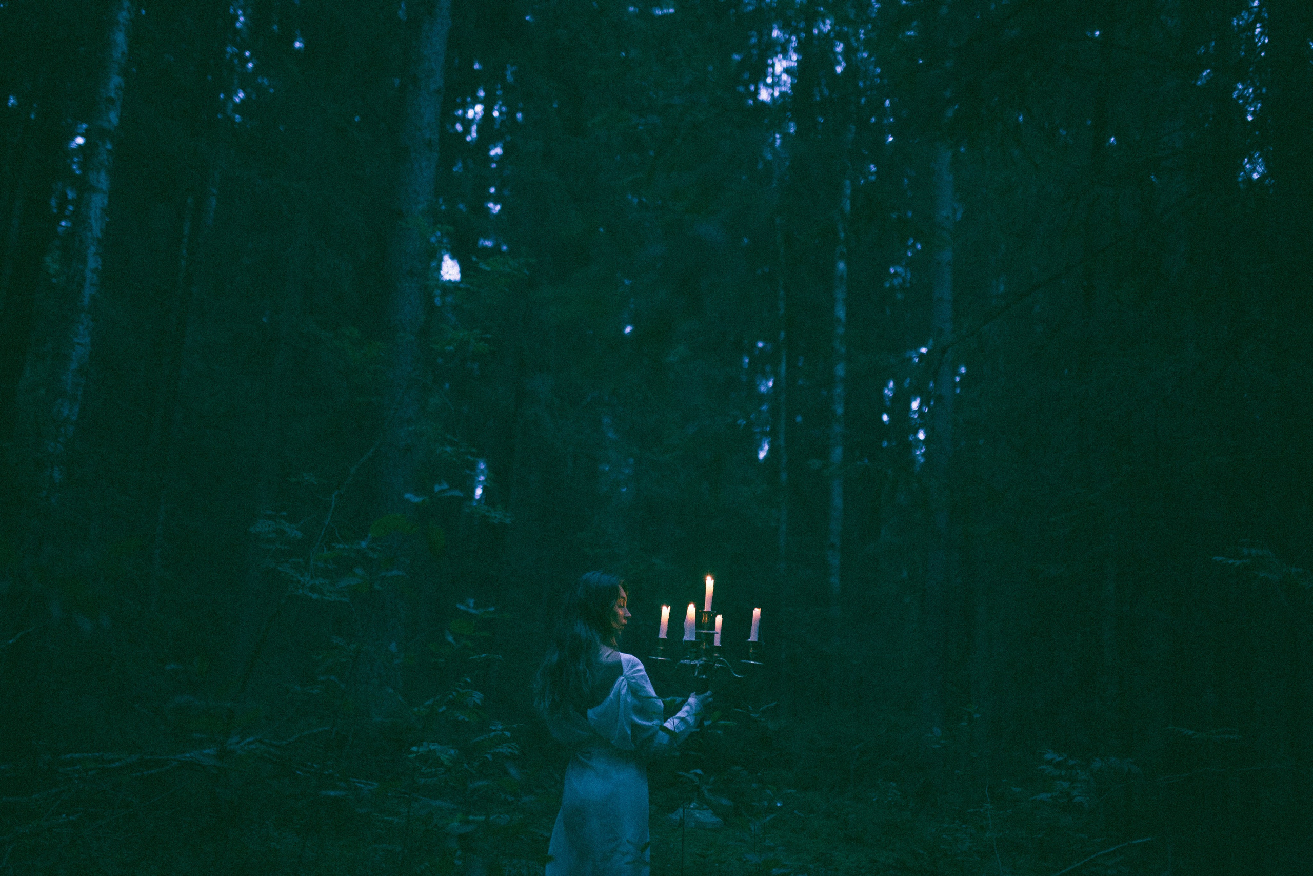 Девушка в белом платье в темном лесу держит канделябр