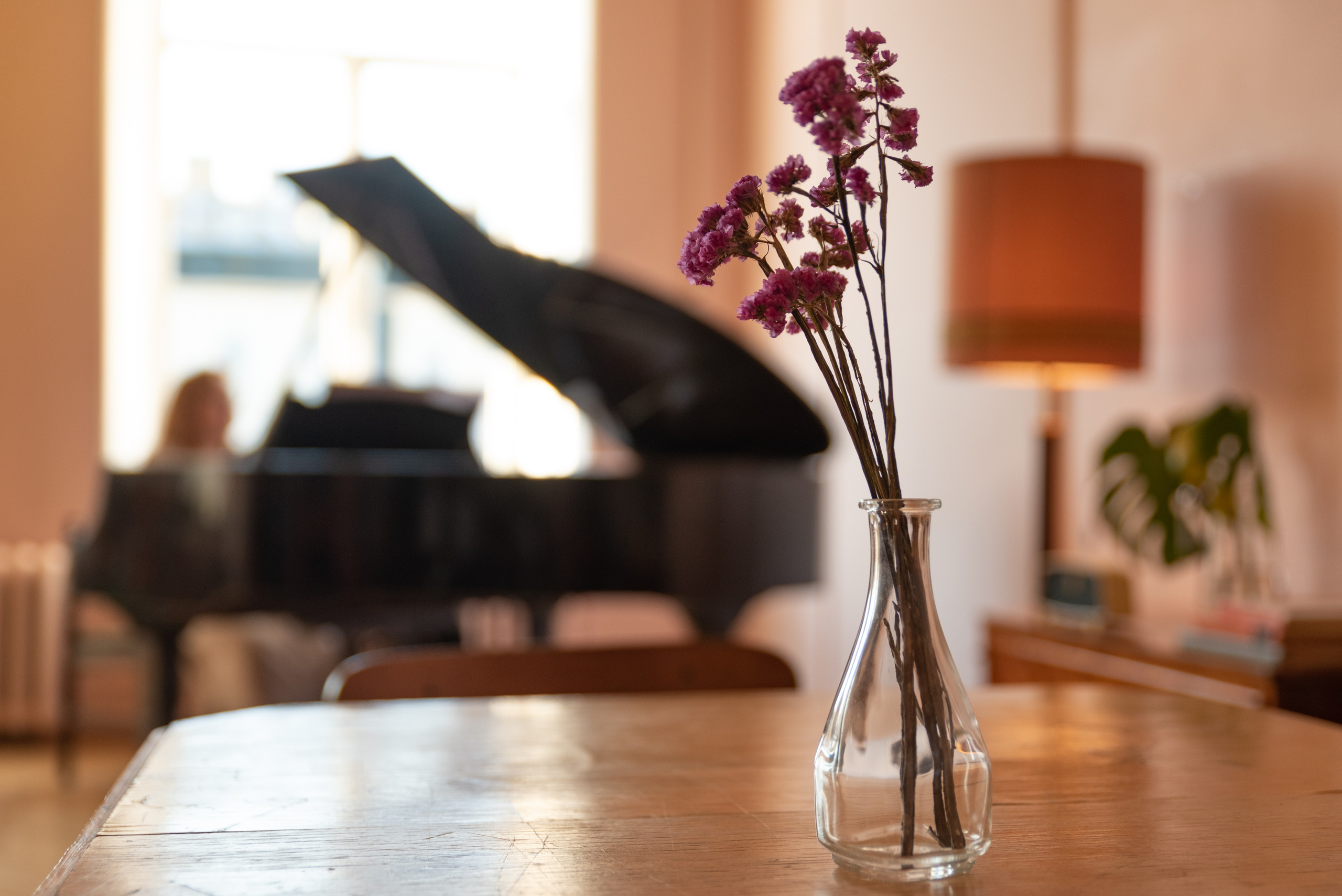 старинный стол, на нём ваза с цветами, на фоне в размытии окно, рояль и музыкант.