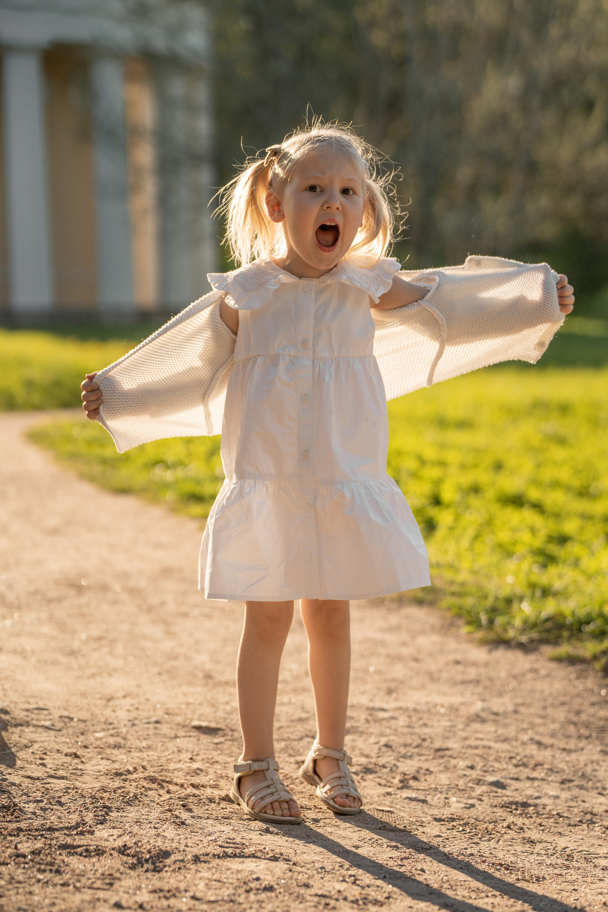 Ребенок кричит в парке и поднялась на носочки. Девочка одета в белое платье и кофточку.