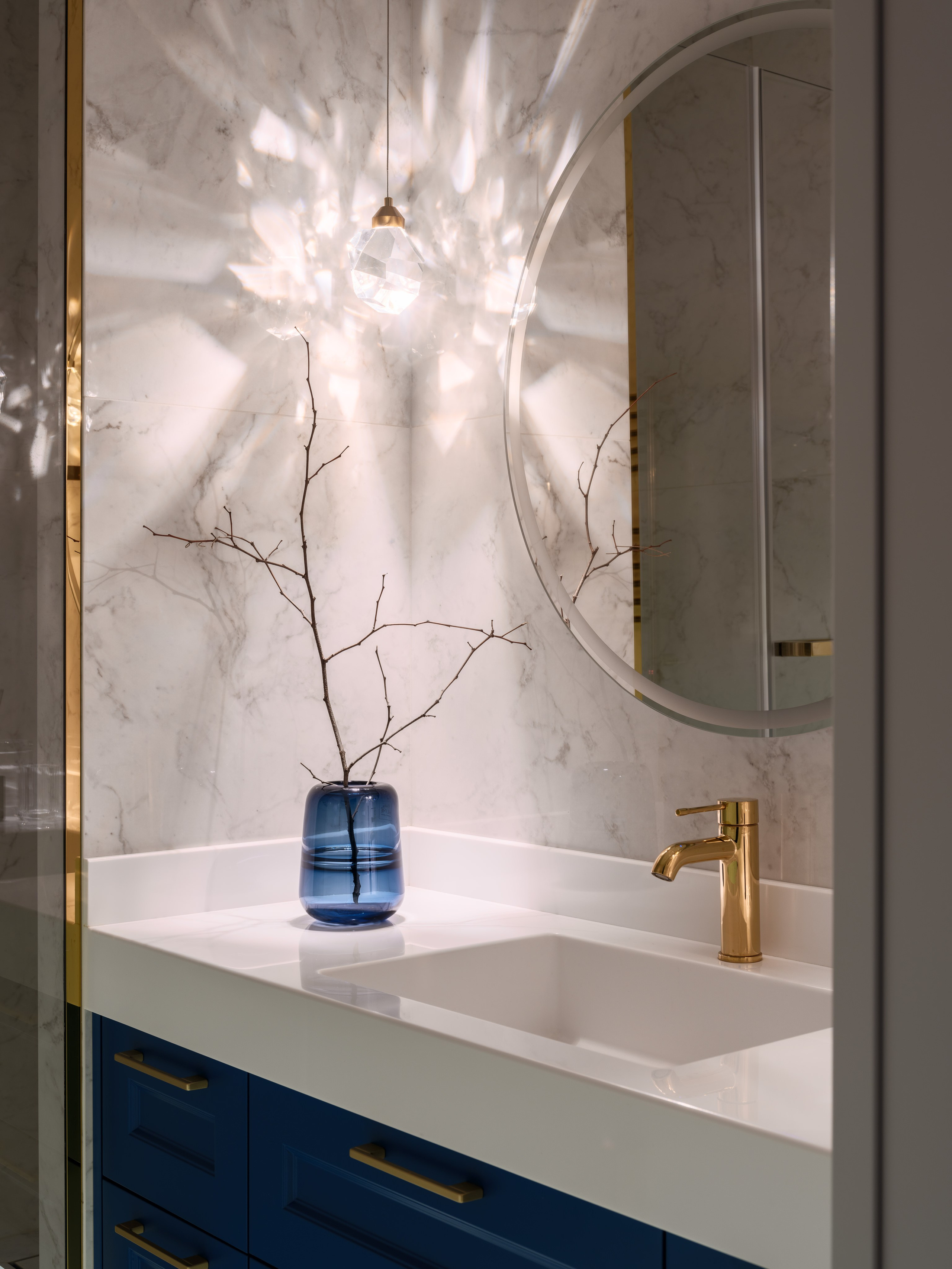 Элегантная ванная комната с круглым зеркалом, подвесным светильником и синим шкафчиком с золотыми ручками. На столешнице белая раковина и ваза с веткой. Интерьер создан дизайнером Евгенией Ермошиной, фото Сергея Денисенко.