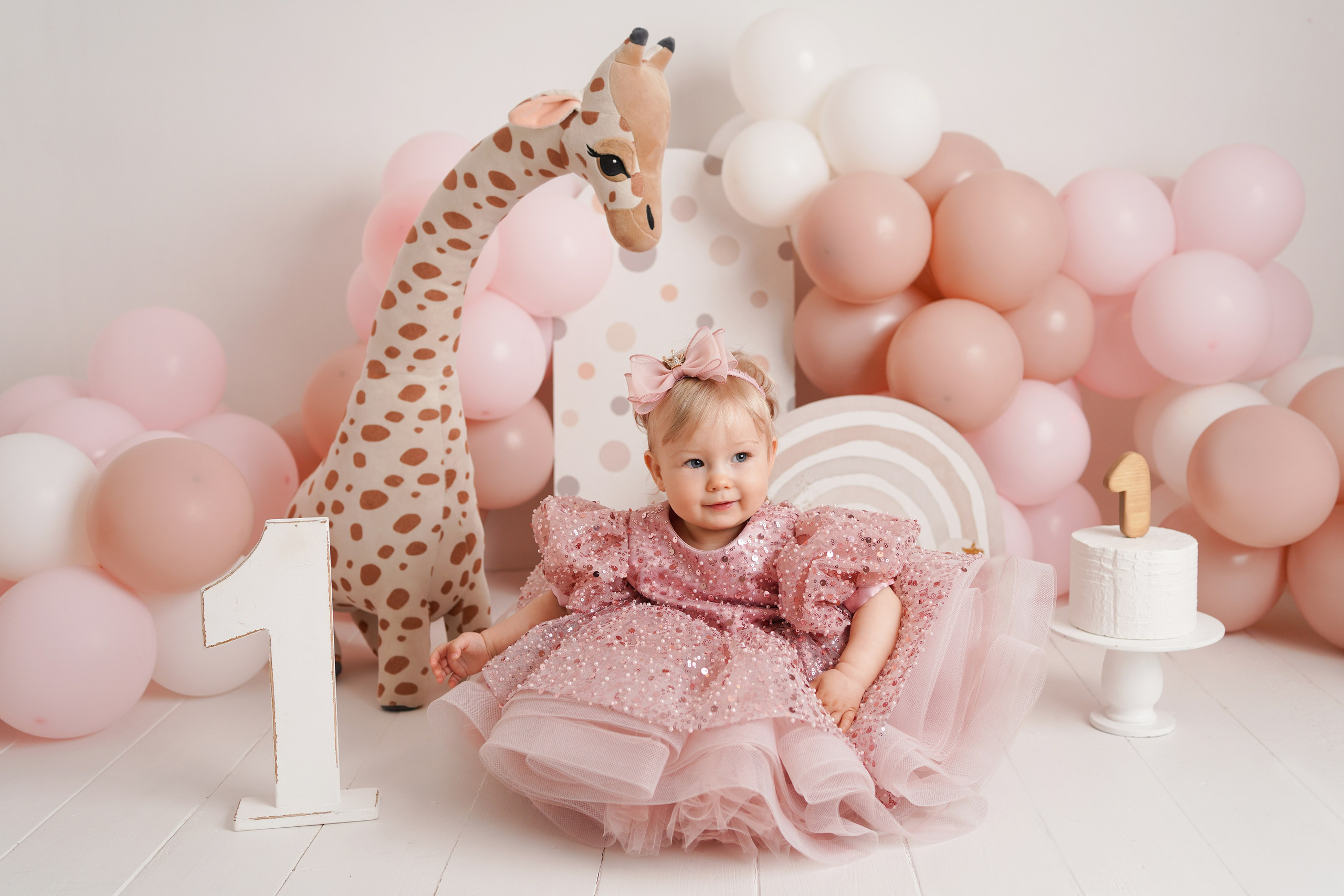 На фото девочка в красивом розовом платье. Рядом жираф, радуга, единица, много воздушных шаров.