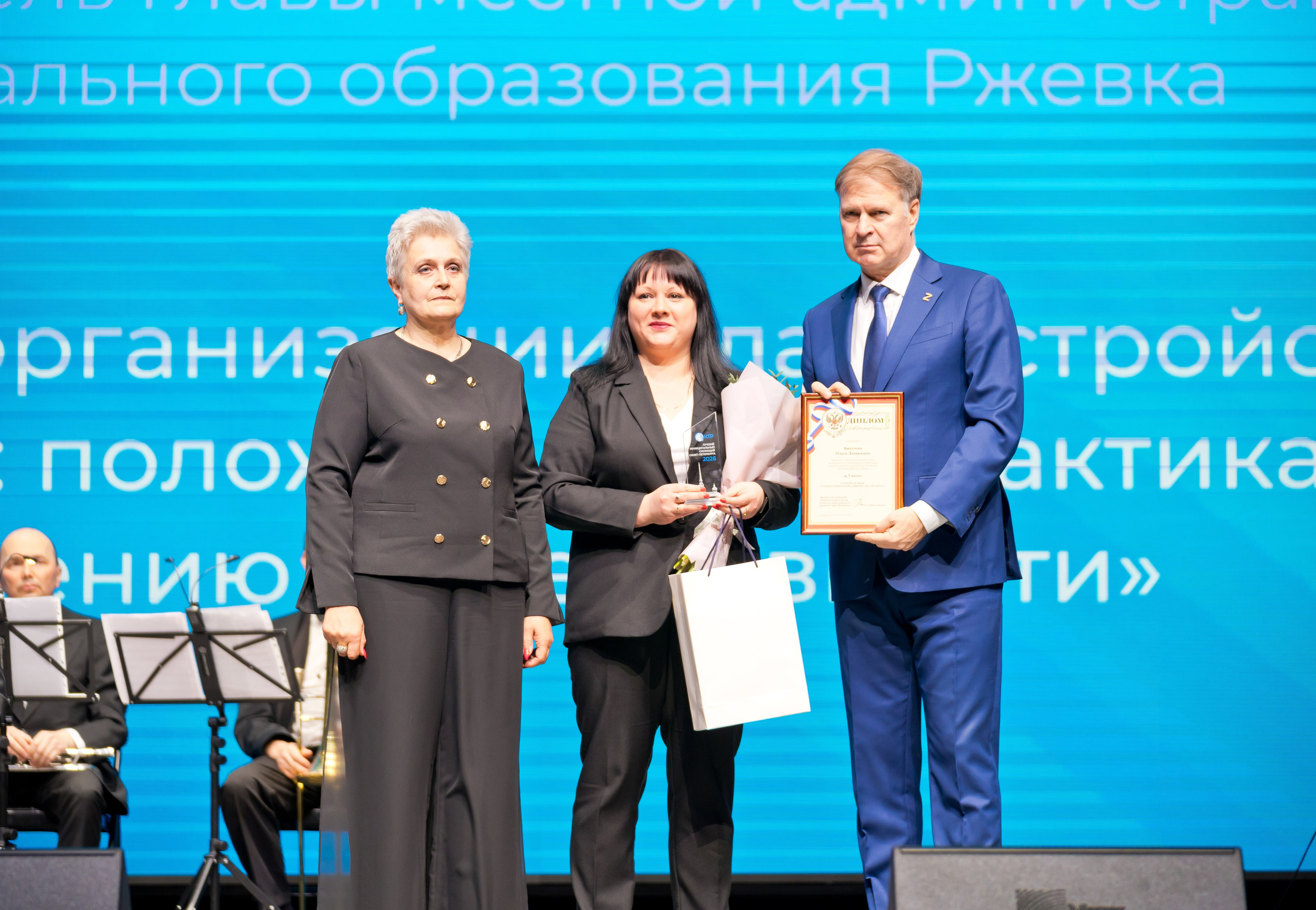 Награждение победителей. Женщина в чёрном костюме, лауреат и глава администрации с грамотой на сцене. День местного самоуправления Санкт-Петербург | Фото: Евгений Мальцев.